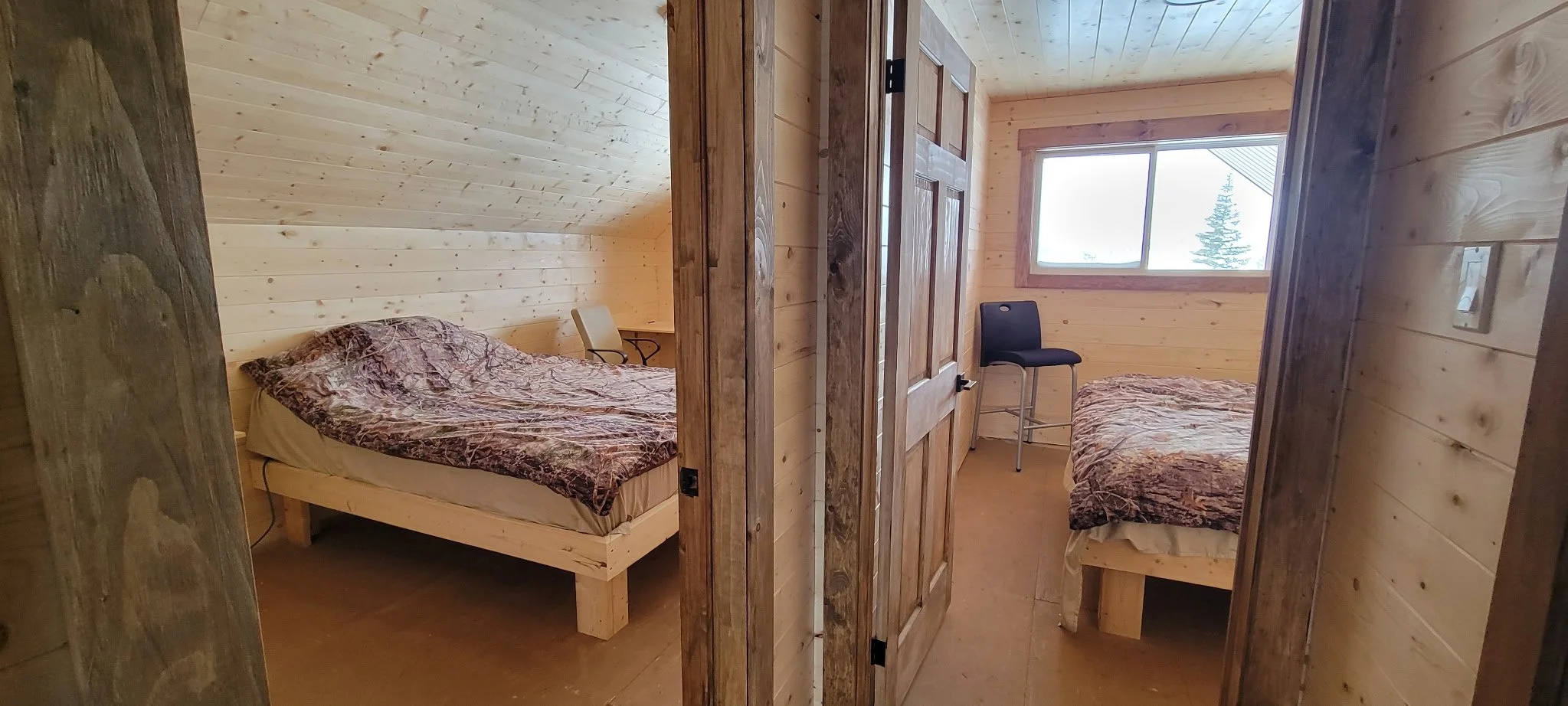 View of two bedrooms in a wooden cabin, each with a bed covered in camouflage-patterned blankets, a chair, and a window revealing trees outside.