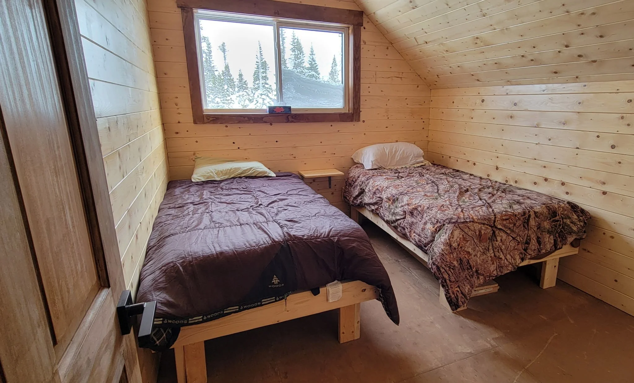 A cozy cabin bedroom with two beds and a window showing snowy trees outside.