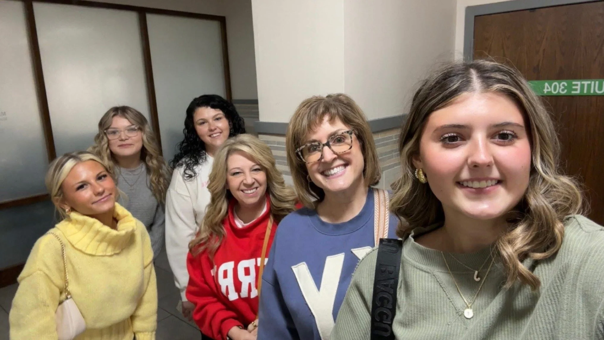 Group of six women taking a selfie in a school hallway, smiling at the camera.