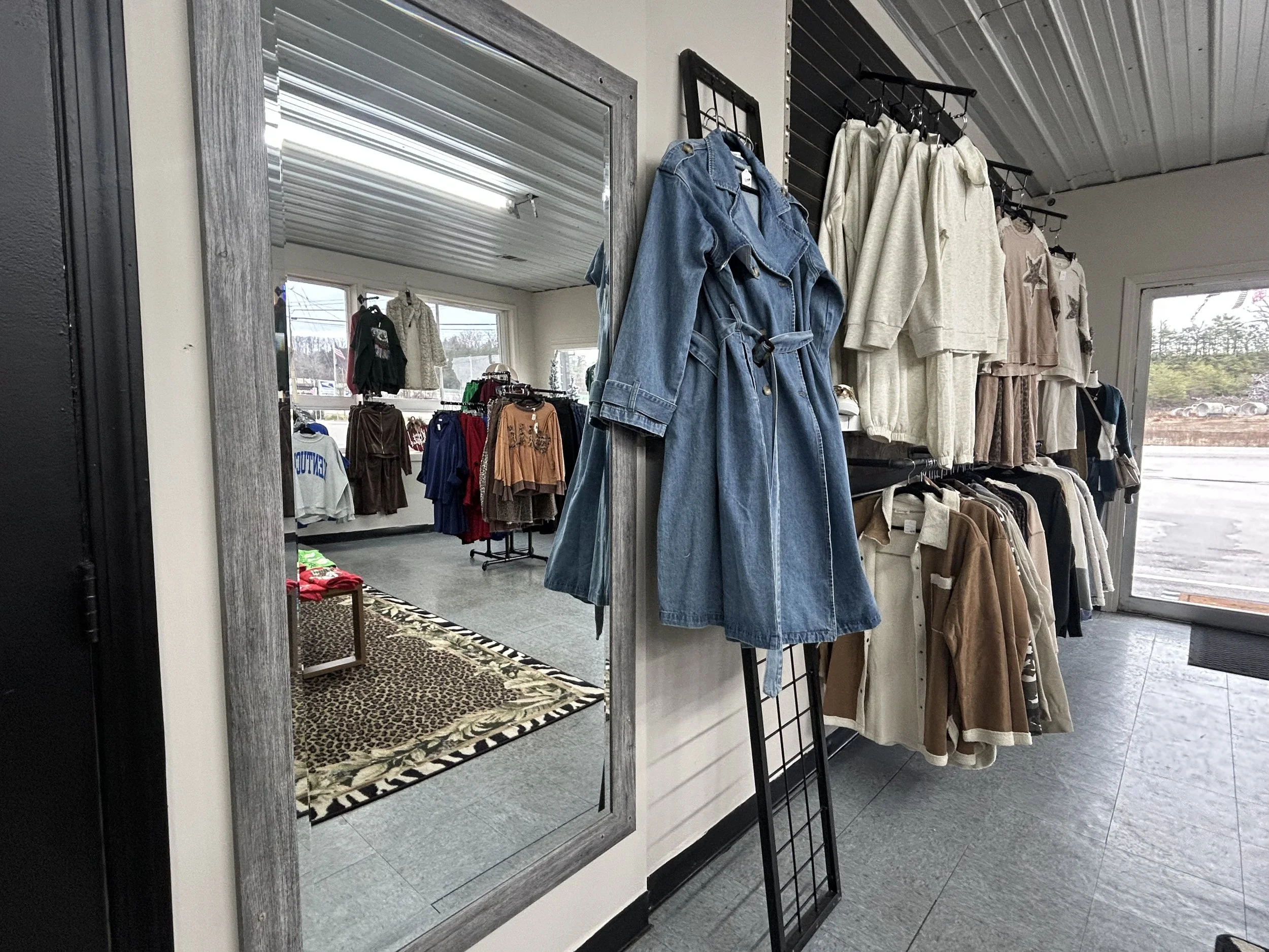 Clothing store display with various jackets and hoodies on racks, a large mirror reflecting more clothing, and a window showing the parking lot outside.