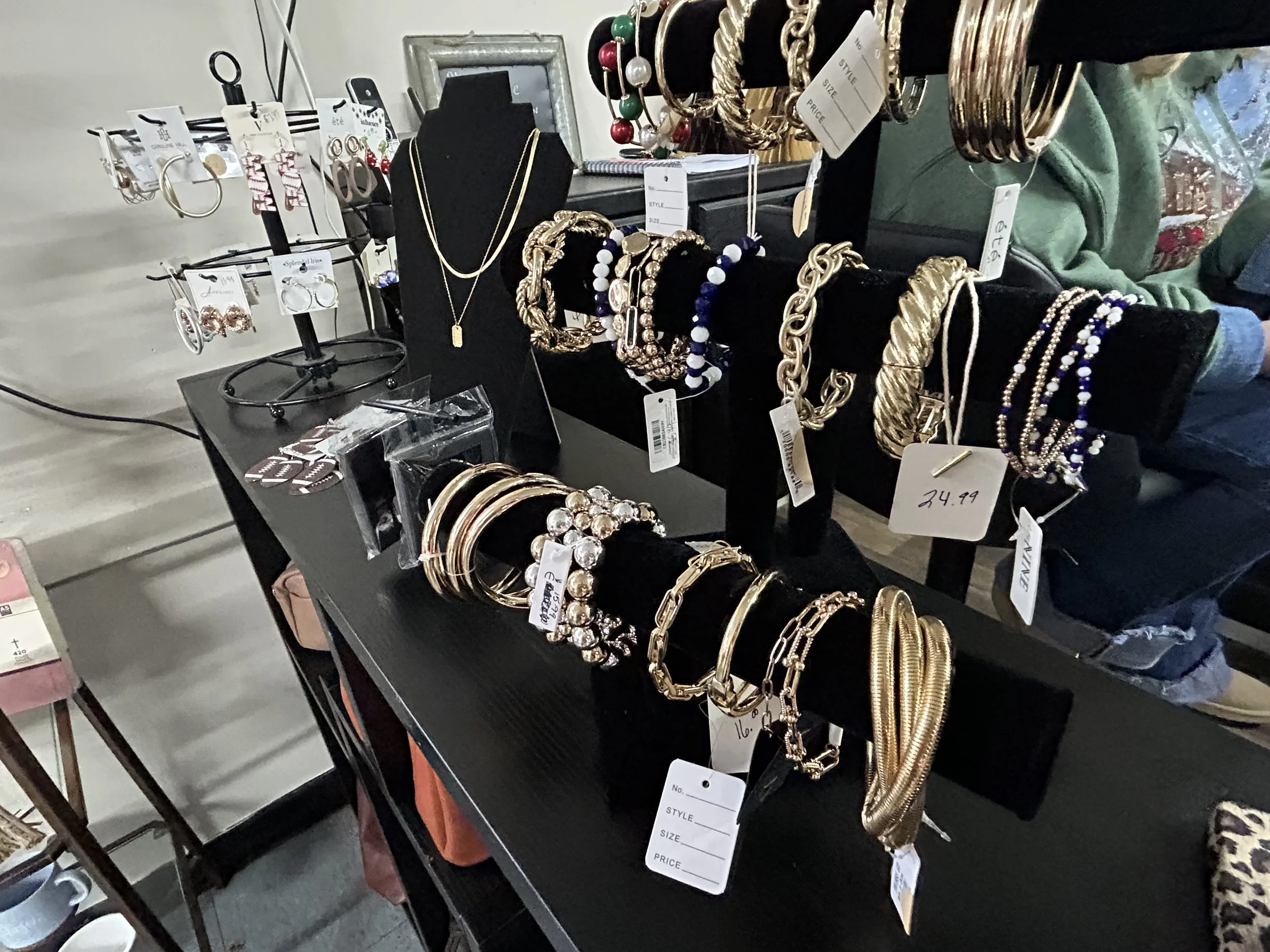 Jewelry display table with gold and silver necklaces, bracelets, earrings, and beaded jewelry on black stands and tags at the shop.
