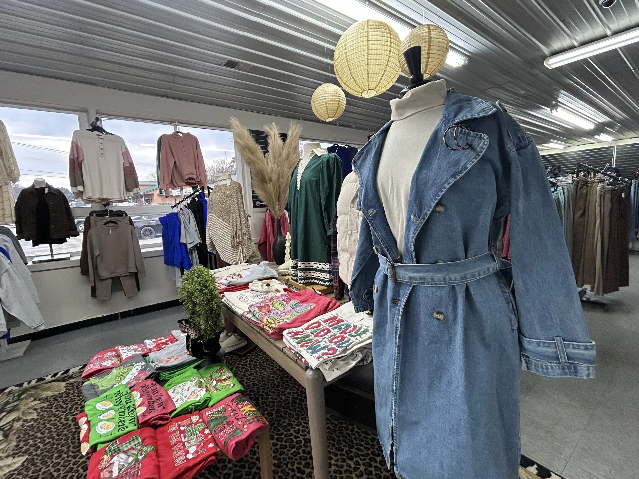 Display of casual clothing including a denim trench coat on a mannequin, surrounded by sweaters, T-shirts, and holiday-themed socks in a retail store with paper lantern decorations.