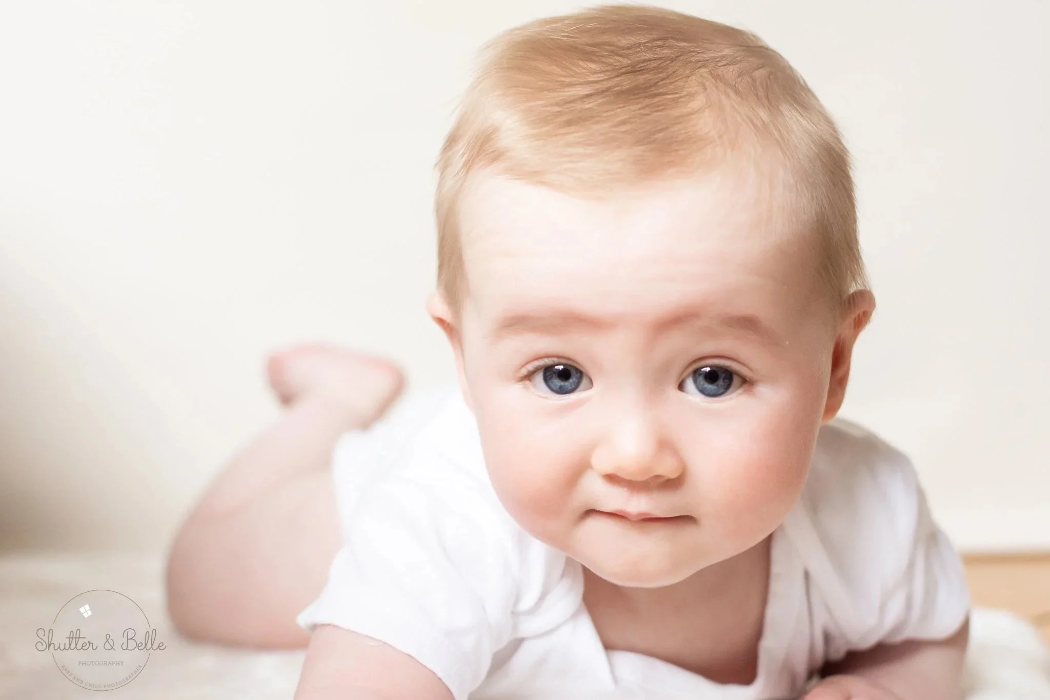 Close-up of a young child with blonde hair and blue eyes, wearing a white shirt, looking directly at the camera while crawling.