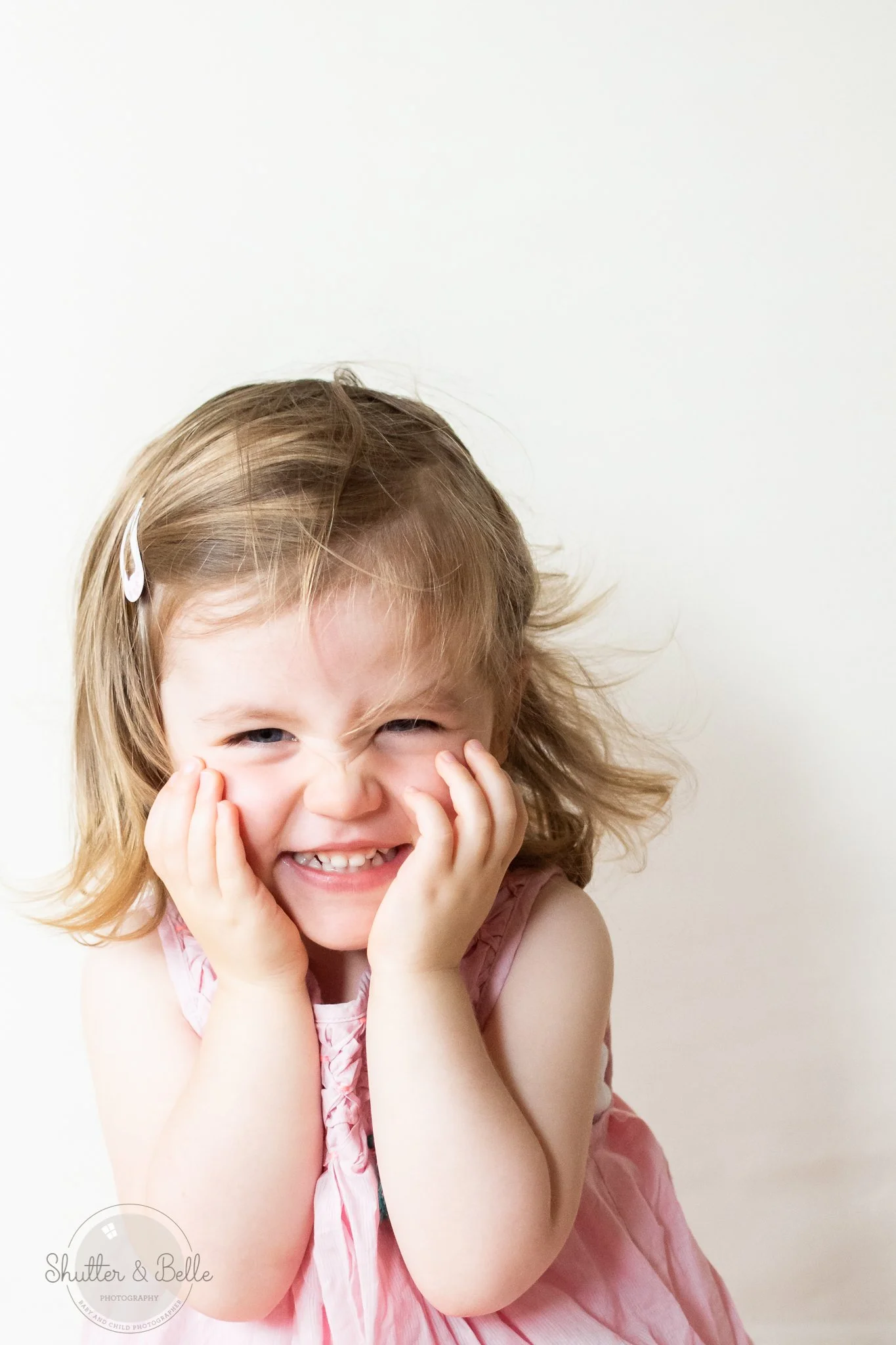 A young girl with light brown hair, wearing a pink dress, is smiling and laughing with her hands on her cheeks against a plain white background.