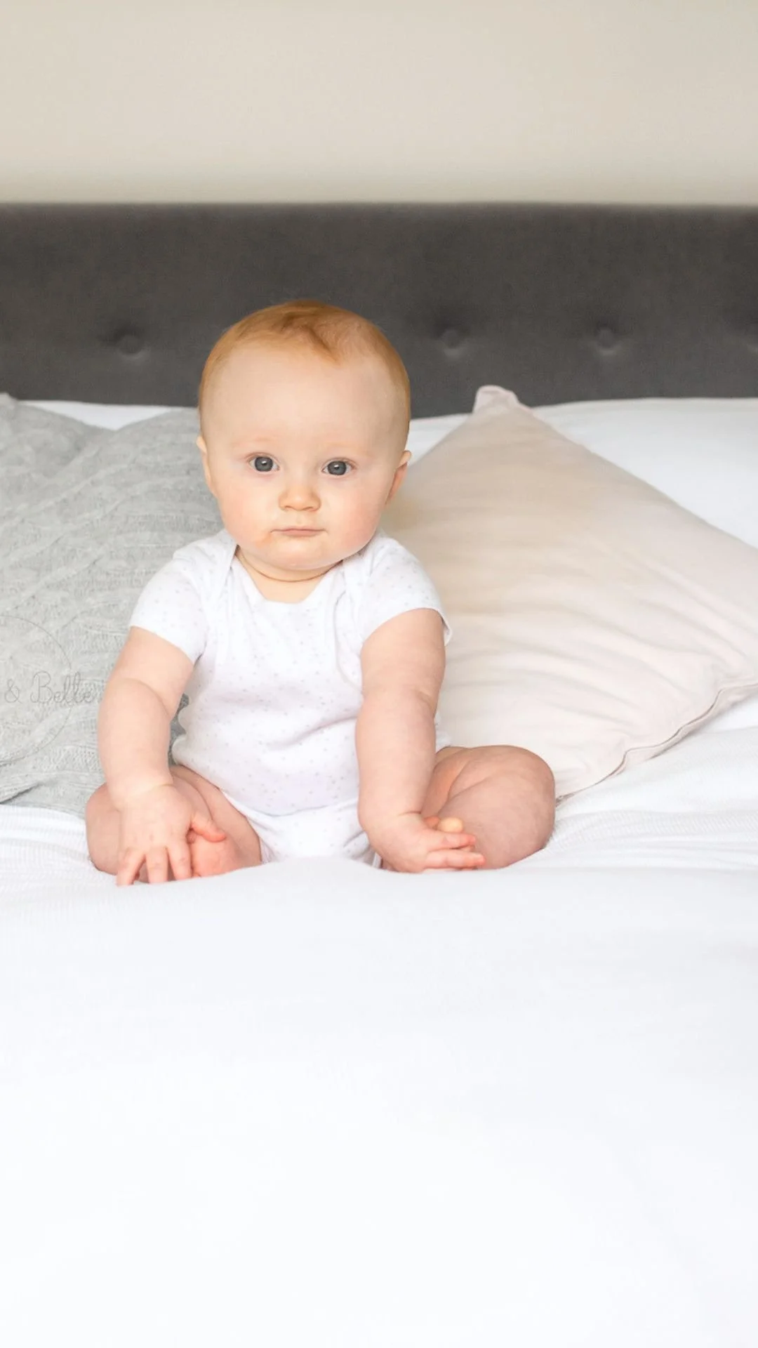 A baby with red hair sitting on a bed with pillows.