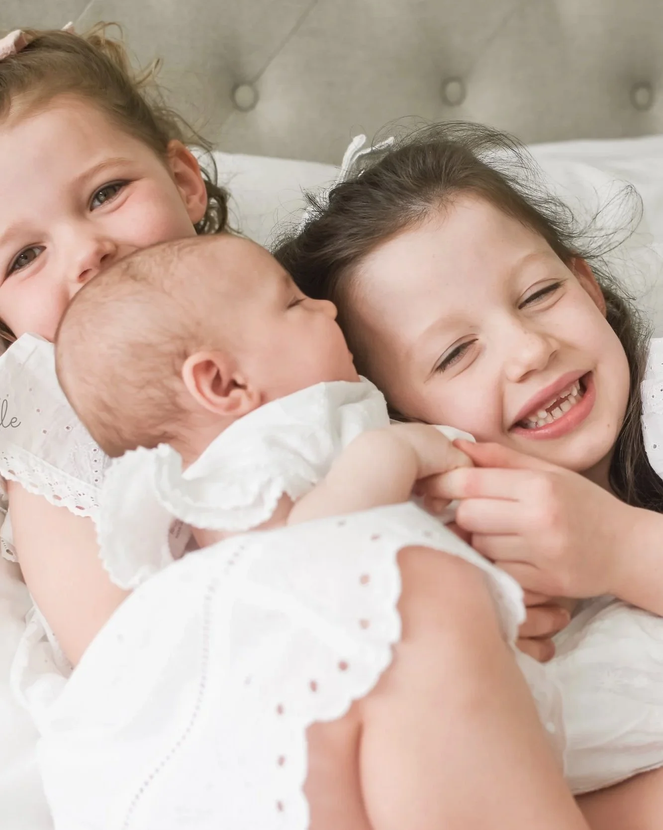 Three young girls, two with brown hair and one with blonde, are cuddling and smiling joyfully while holding a newborn baby.