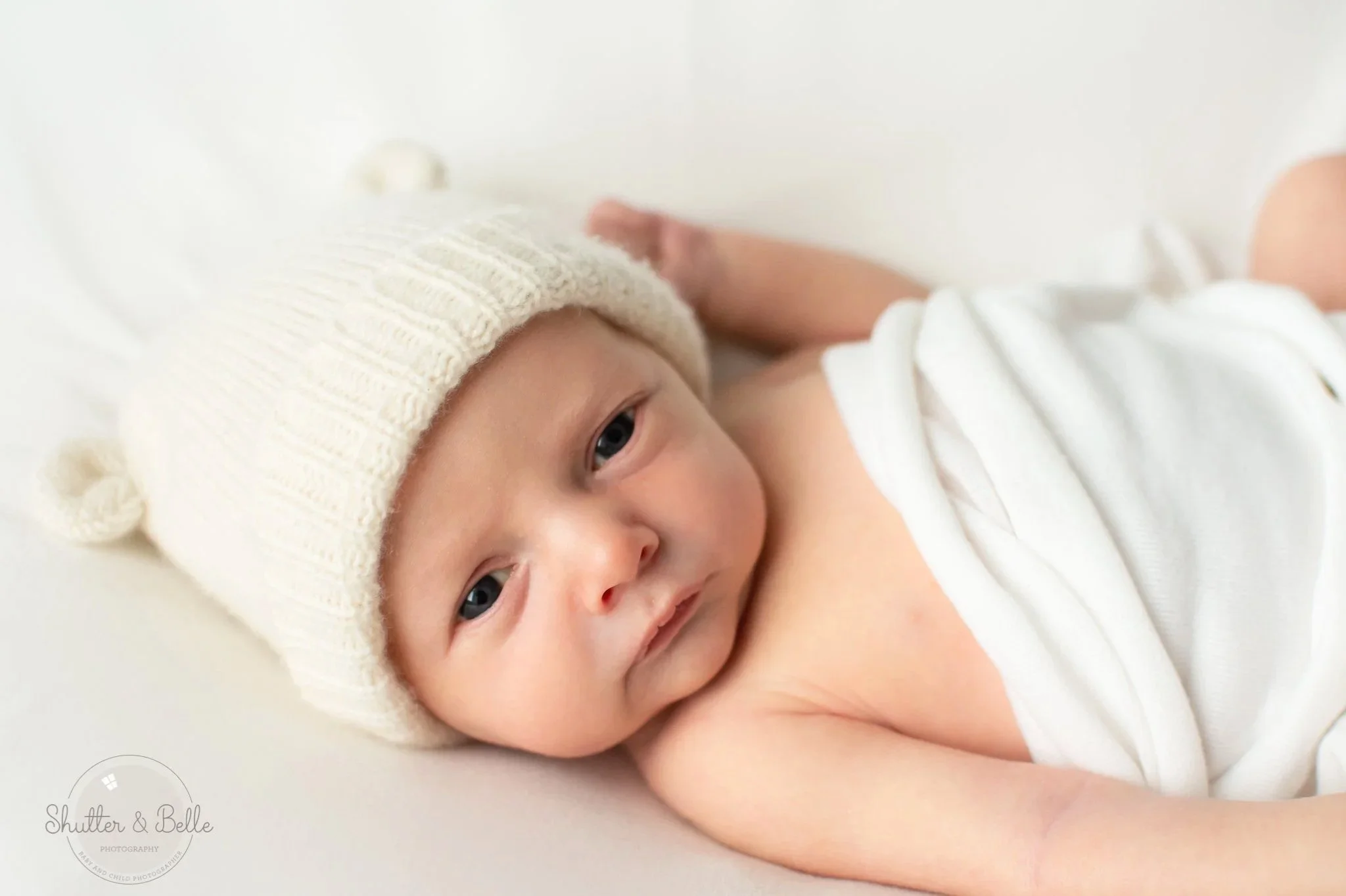 Close-up of a baby lying on white surface, wearing a knitted cream hat, with blue eyes looking towards camera, smiling slightly.