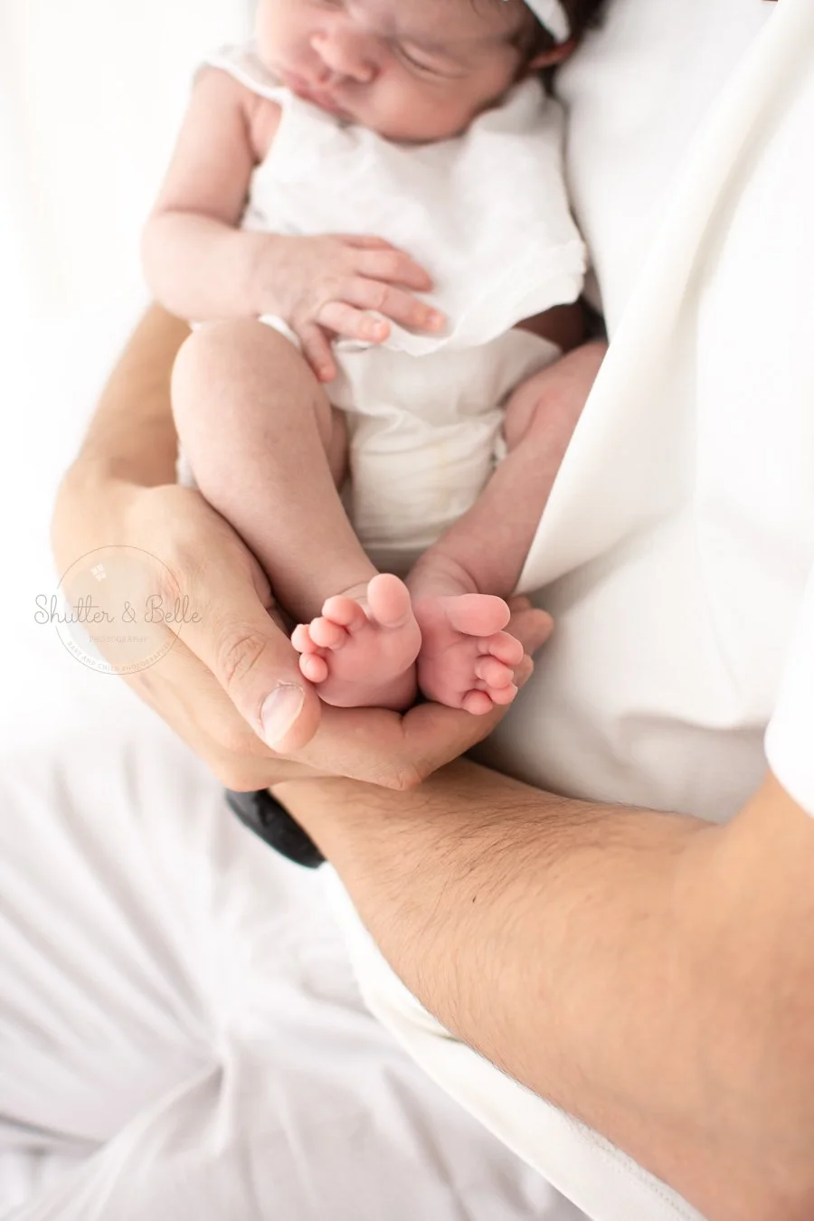 Close-up of an adult's hand holding a newborn baby with tiny feet, the baby is sleeping and wearing a white outfit, with a soft white background.