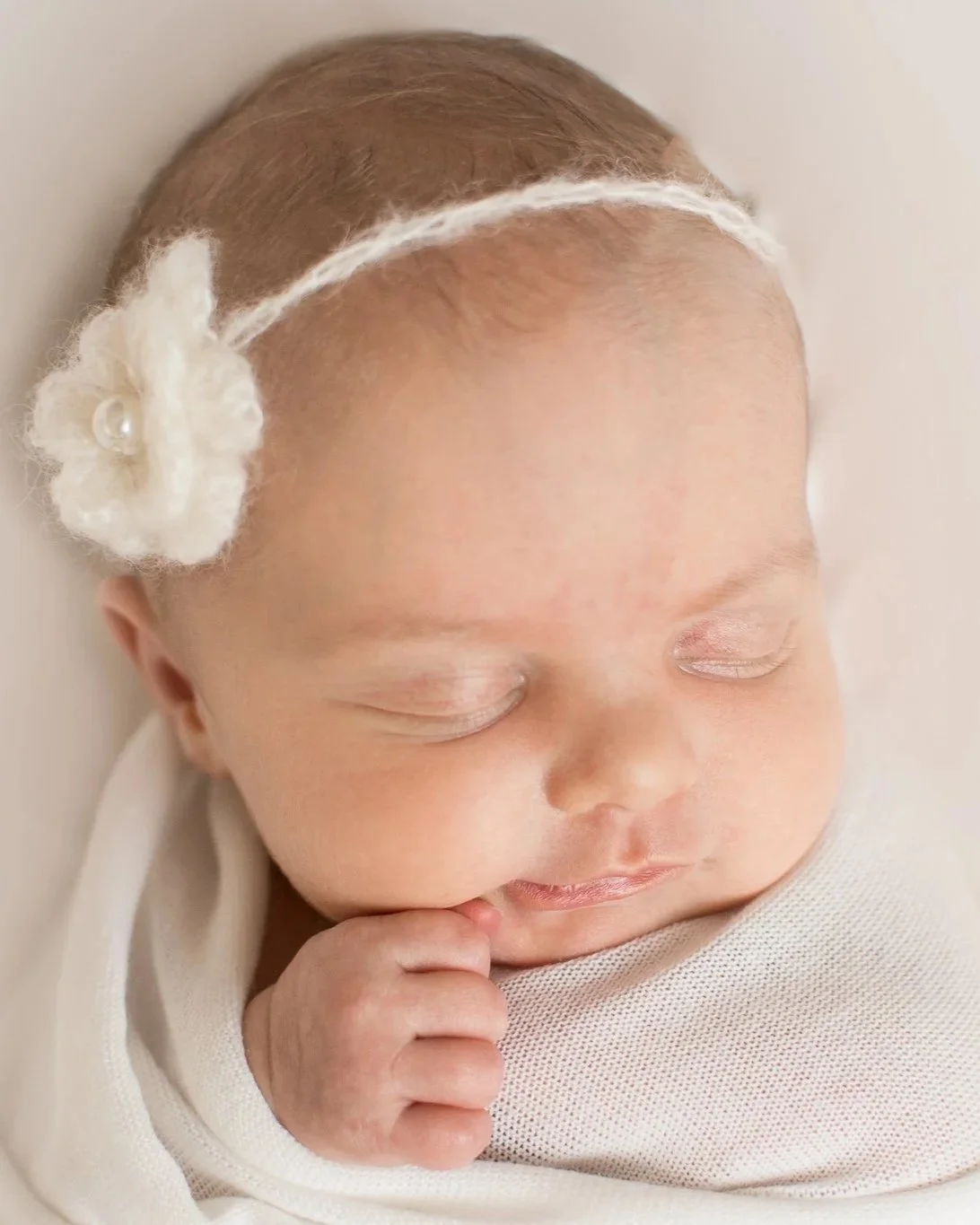 Close-up of a sleeping newborn baby with a floral headband and light-colored clothing.