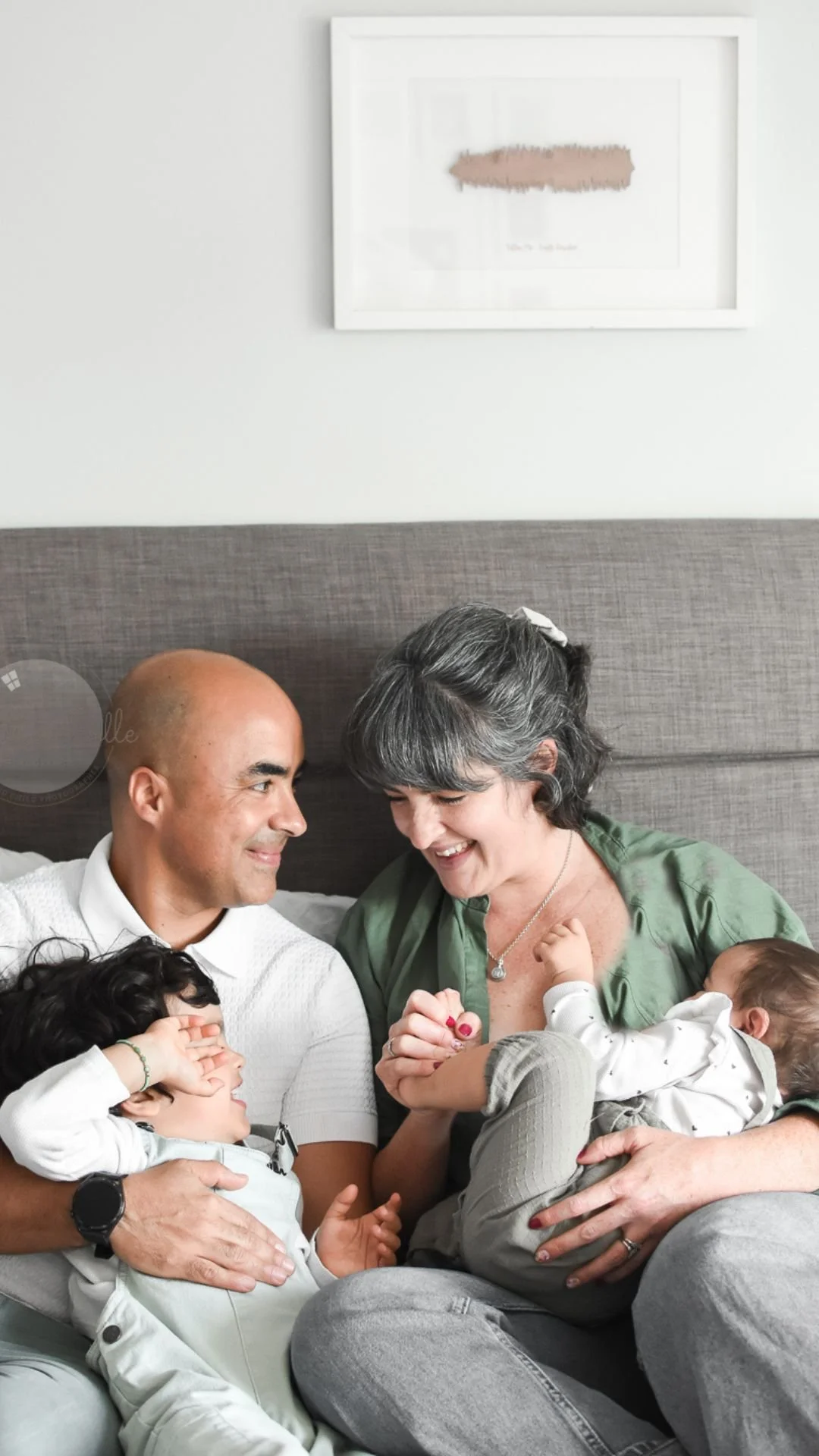 A family of four, including a bald man, an older woman with gray hair, a young boy, and a baby, sitting on a bed at home smiling and playing together.