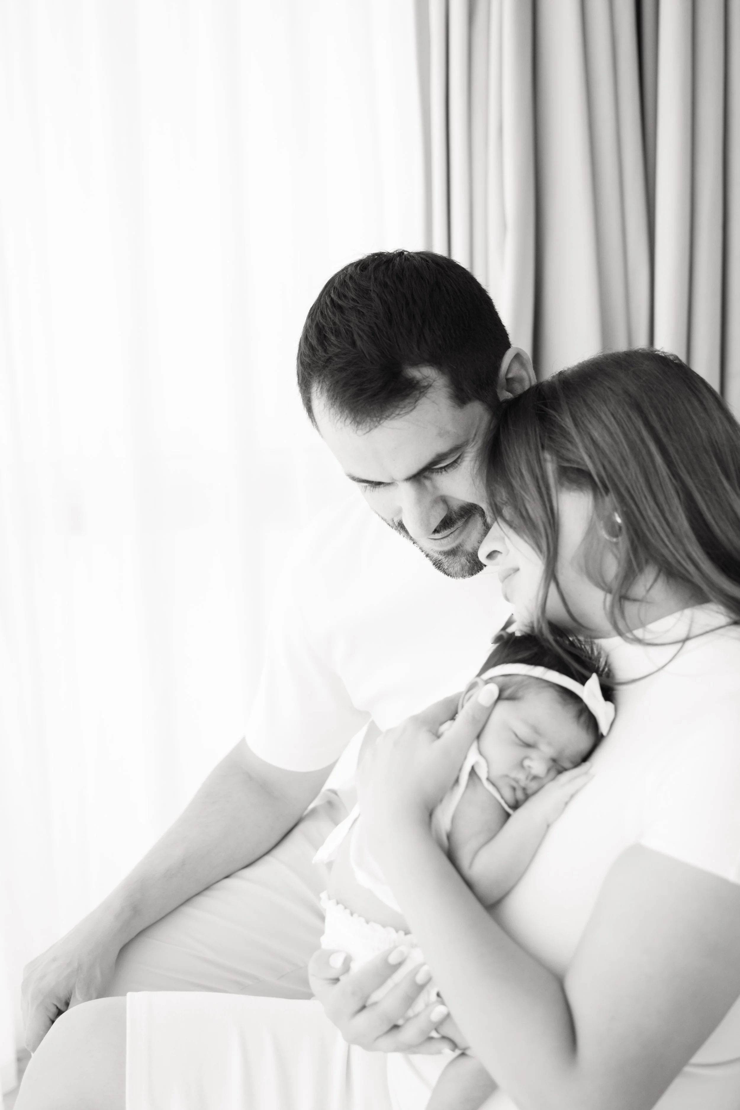 A black and white photo of a man, woman, and baby. The man and woman are embracing and holding the sleeping baby, who is nestled in the woman's arms. The background features curtain drapes.