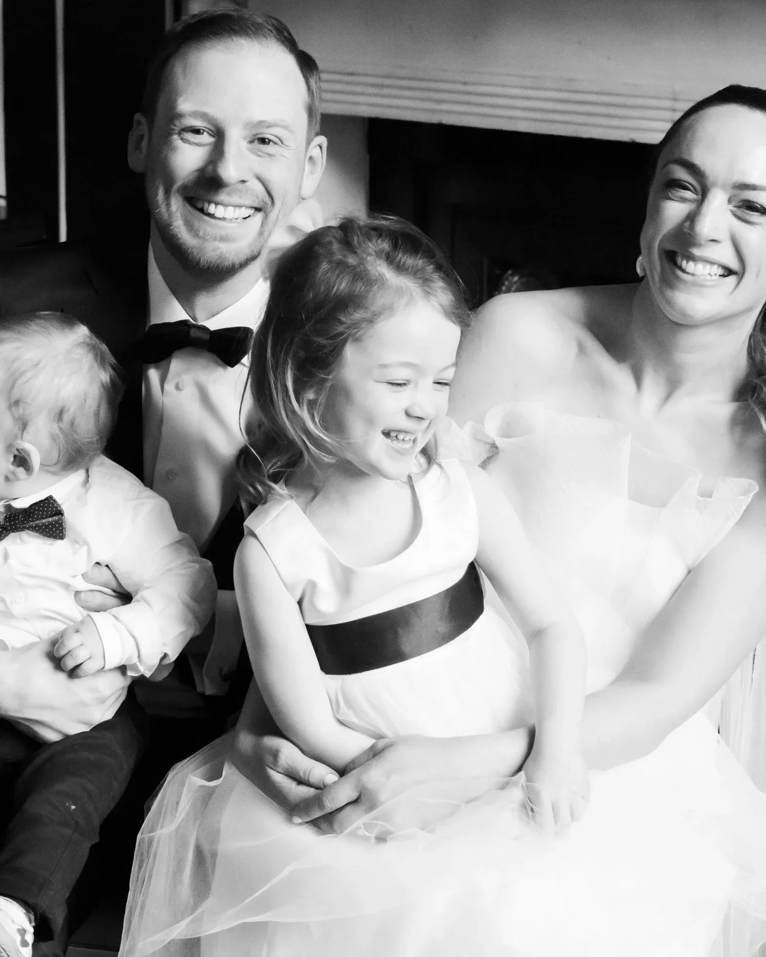 Black and white photo of a smiling man in a tuxedo with a bowtie, holding a young girl in a white dress with a dark waistband, both smiling, with two other children partially visible.