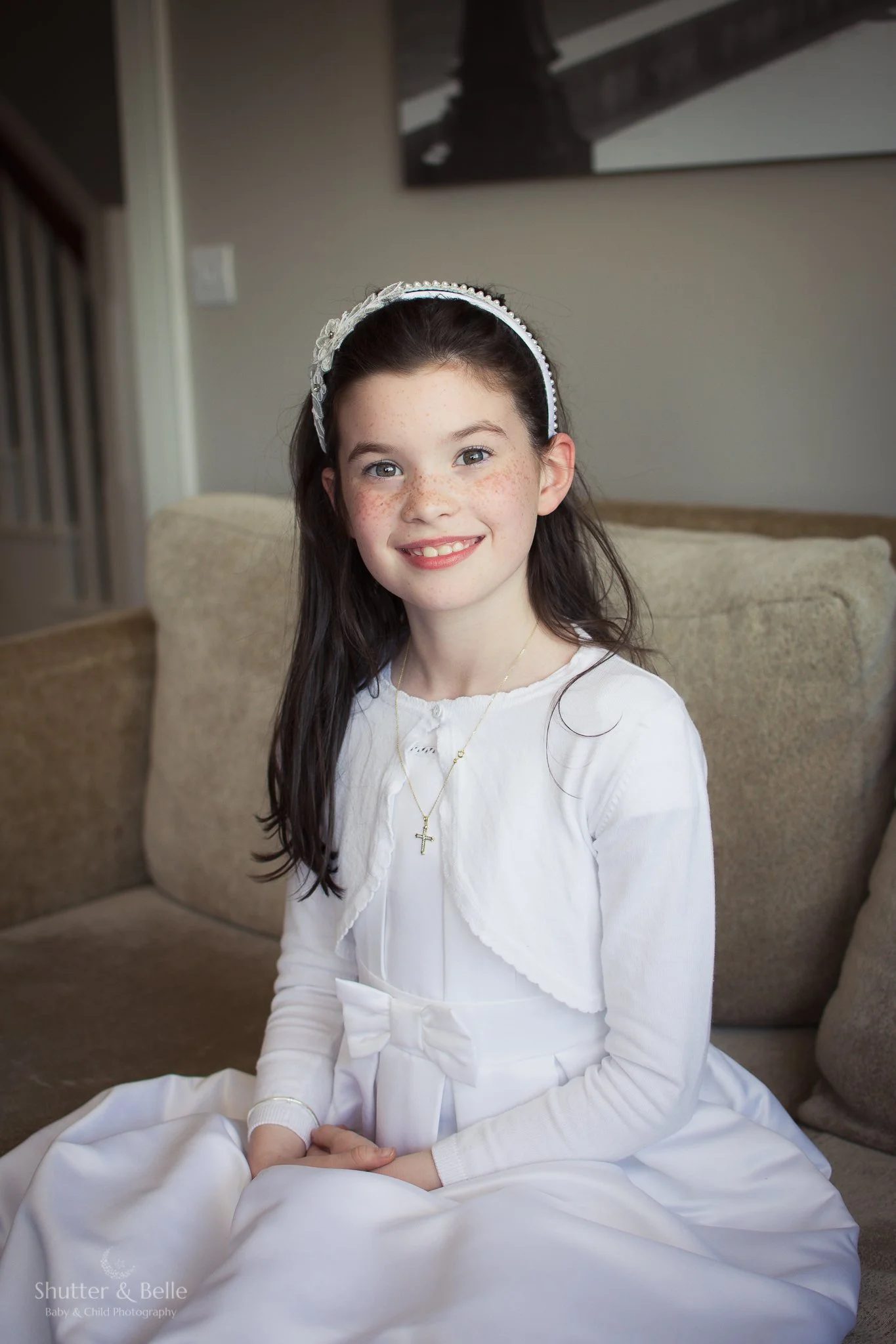 A young girl with dark brown hair, wearing a white dress and a white headband, sitting on a beige sofa inside a home with a neutral-colored wall and a piece of black-and-white art in the background.