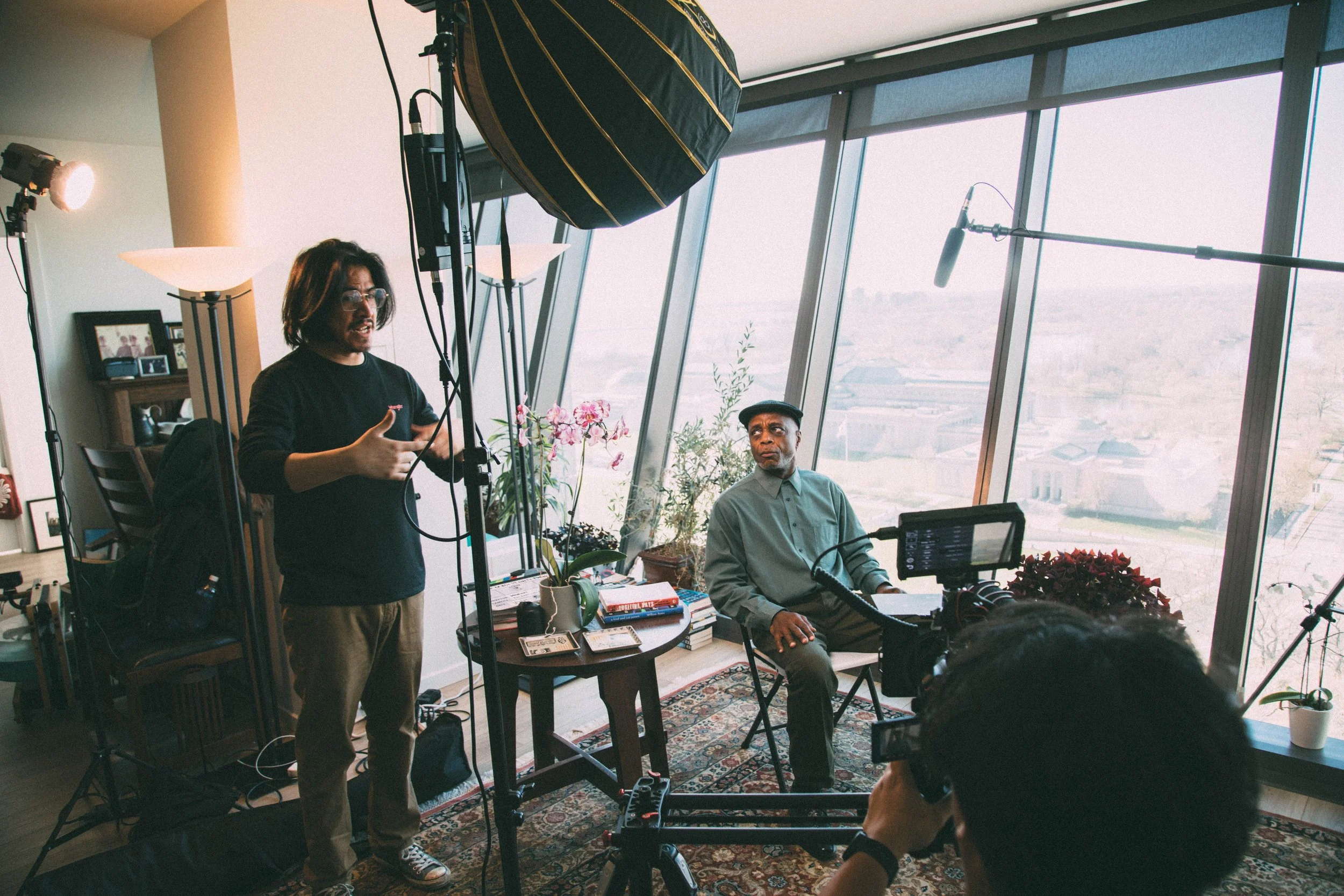 A film or video production crew working indoors near large windows with a city view. One person is standing and speaking, while another is seated, operating equipment. There is a professional camera, lighting, and a boom microphone visible.