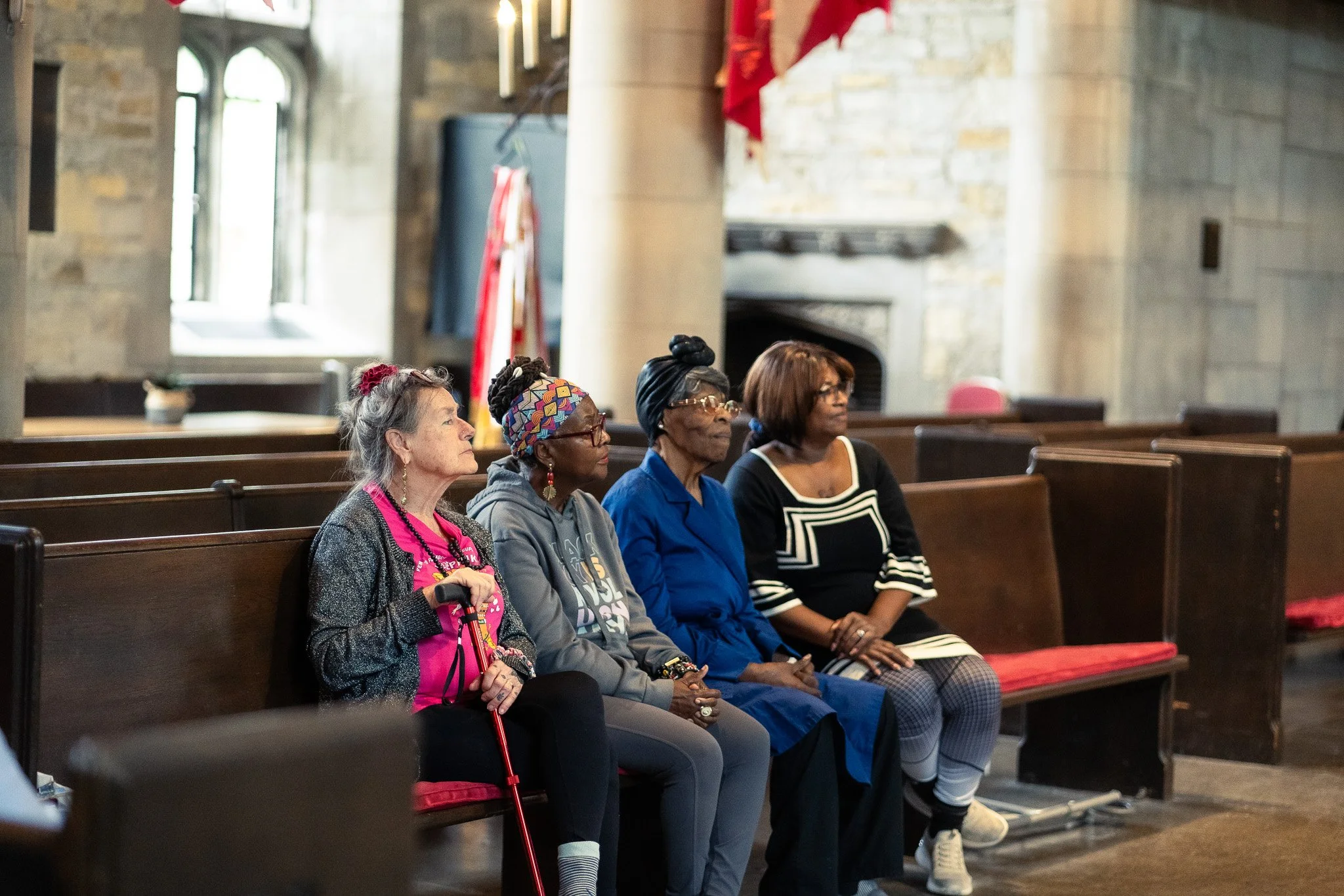 Four women sitting on pews inside a church, attentively listening. The church has stone walls, large windows, and a fireplace in the background.