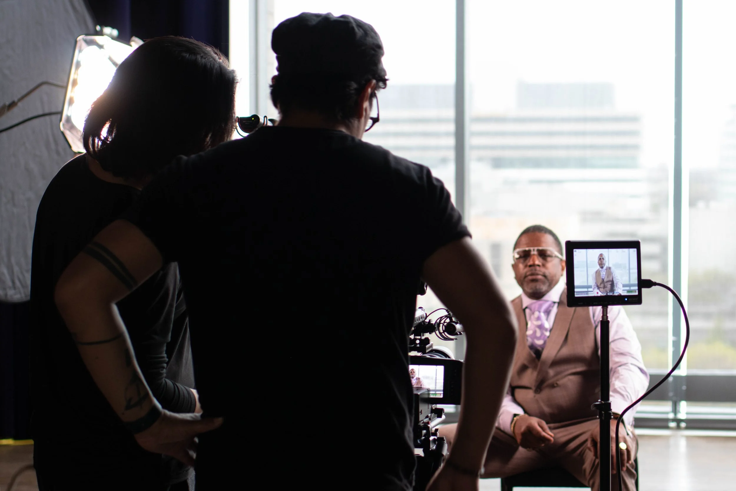 Ronald "Ronnie" Kitchen sitting in front of a camera setup during a video shoot, with crew members behind the camera, and a large window with city buildings in the background.