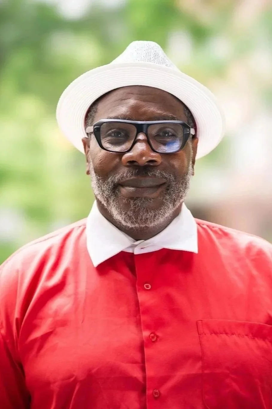 A man with gray beard and glasses wearing a white hat and red shirt, outdoors with blurred green background.