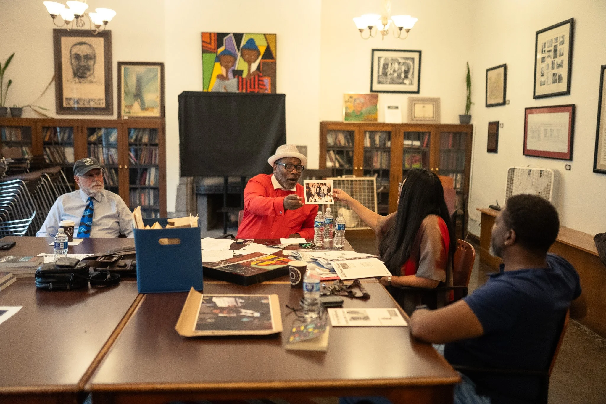 A group of four diverse people sitting around a large wooden table in a room decorated with framed artwork and bookshelves. One man wearing a red shirt, fedora, and glasses, hands a photograph to a woman with long dark hair. The other man, wearing a white shirt and tie, observes. There are water bottles and various papers and magazines on the table.