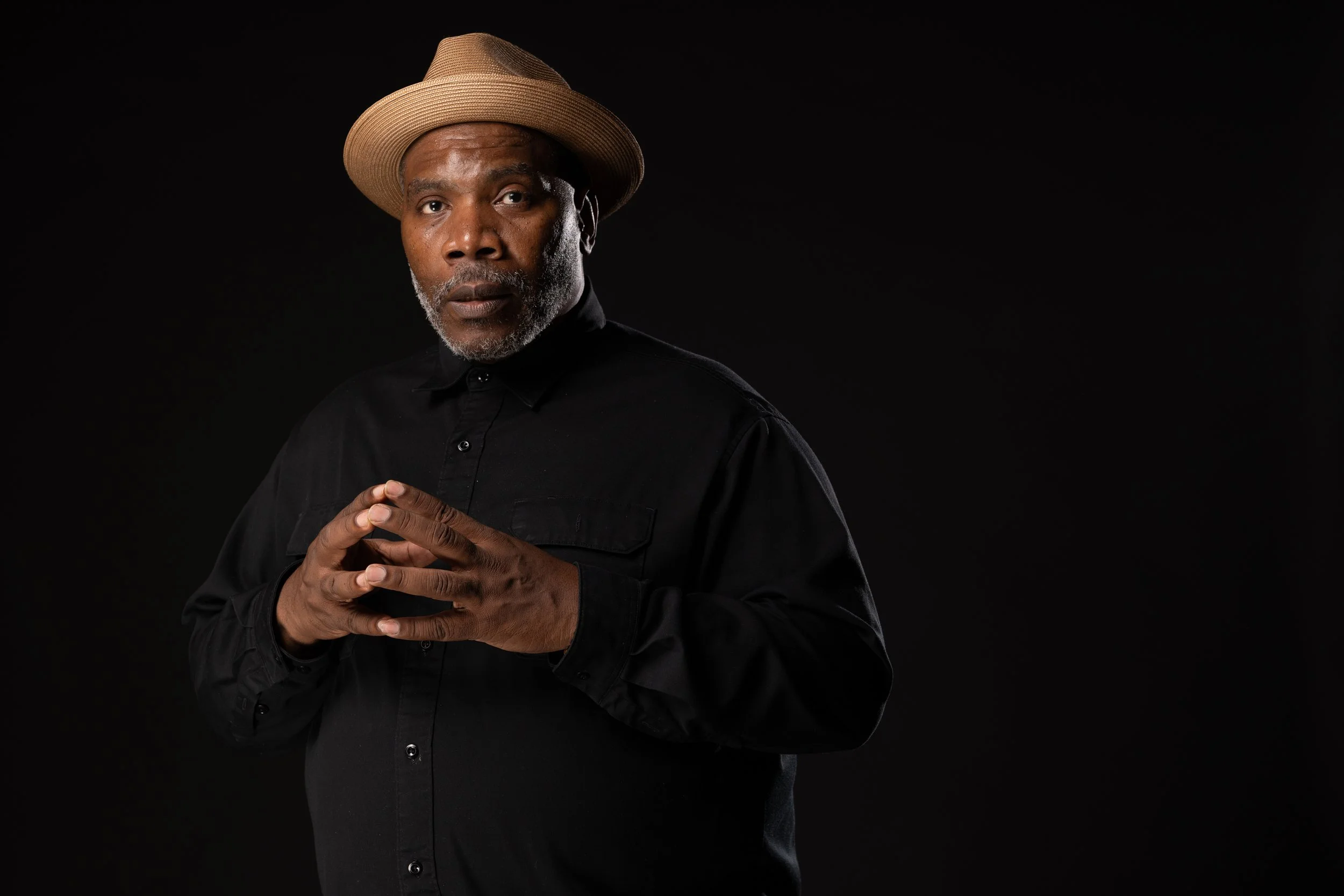 Renaldo Hudson wearing a black shirt and beige fedora, standing against a black background, with his hands clasped together in front of him.