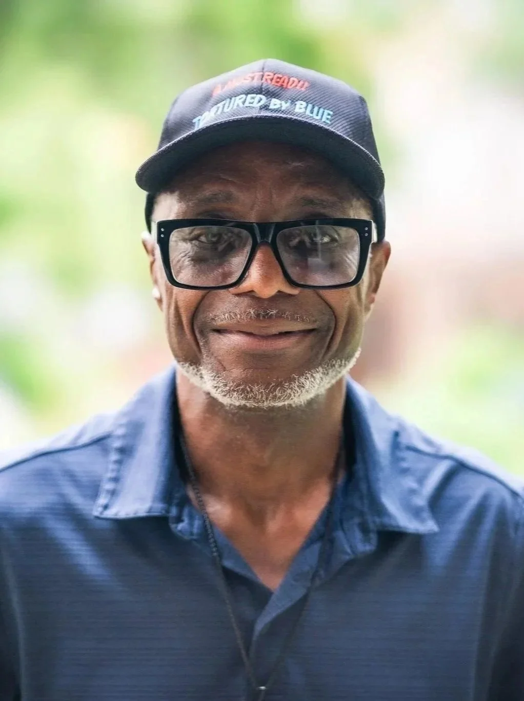 A smiling middle-aged man with glasses and a cap, standing outdoors with a blurred green background.