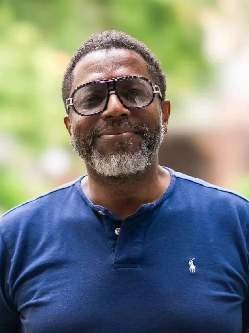 A middle-aged man with gray beard wearing large sunglasses and a navy blue Polo Ralph Lauren shirt, smiling outdoors with a green, blurred background.