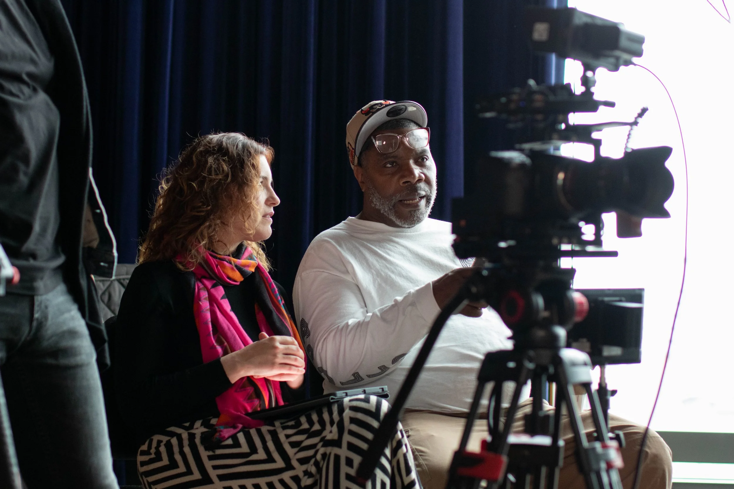 Renaldo Hudson and Elizabeth Futrell seated behind a professional video camera, engaged in a discussion or interview in a dark room with a bright light source behind them.