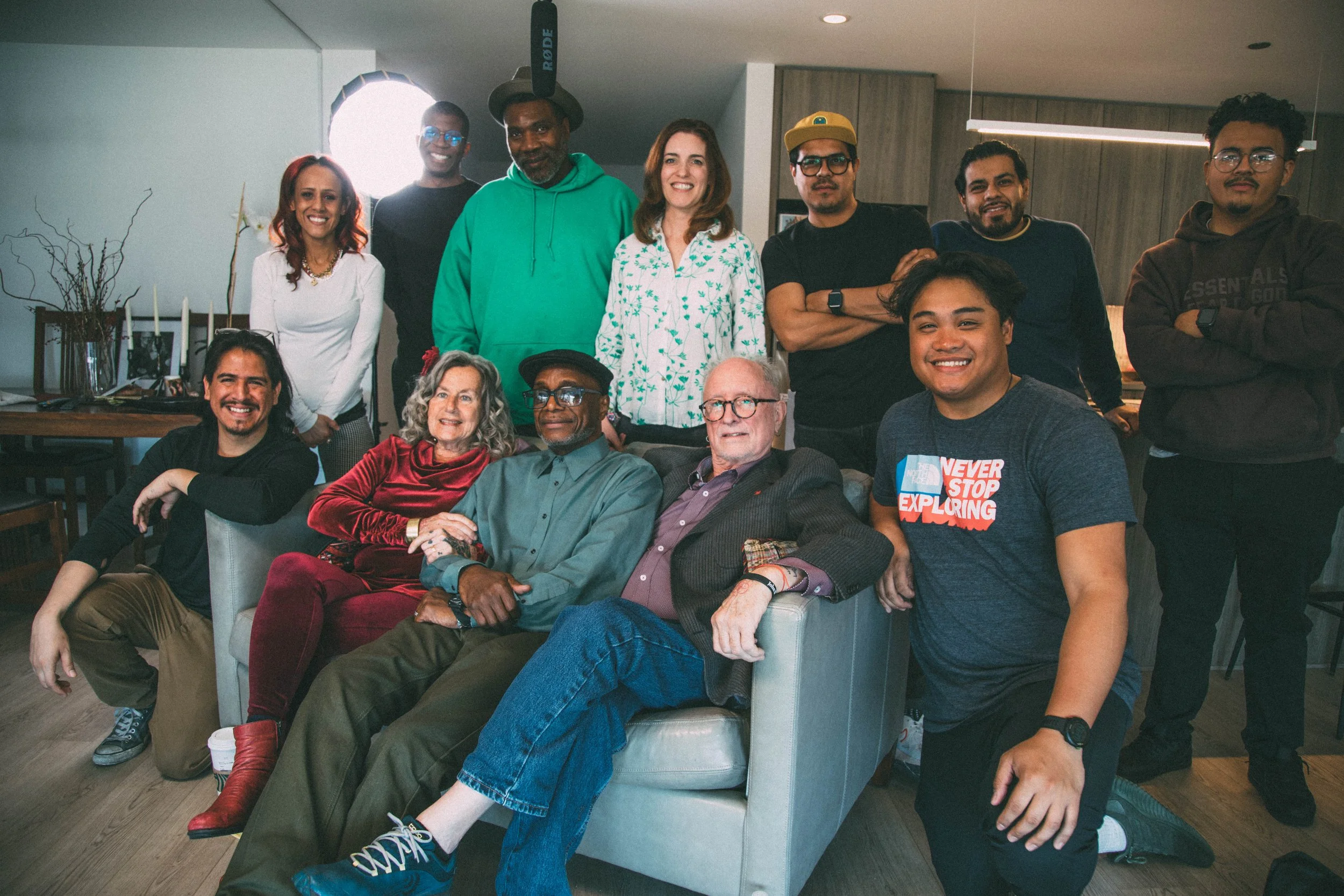 A diverse group of fifteen people posing for a photo in a modern indoor setting, with some seated on a sofa and others standing behind, showing a variety of ages, ethnicities, and styles.