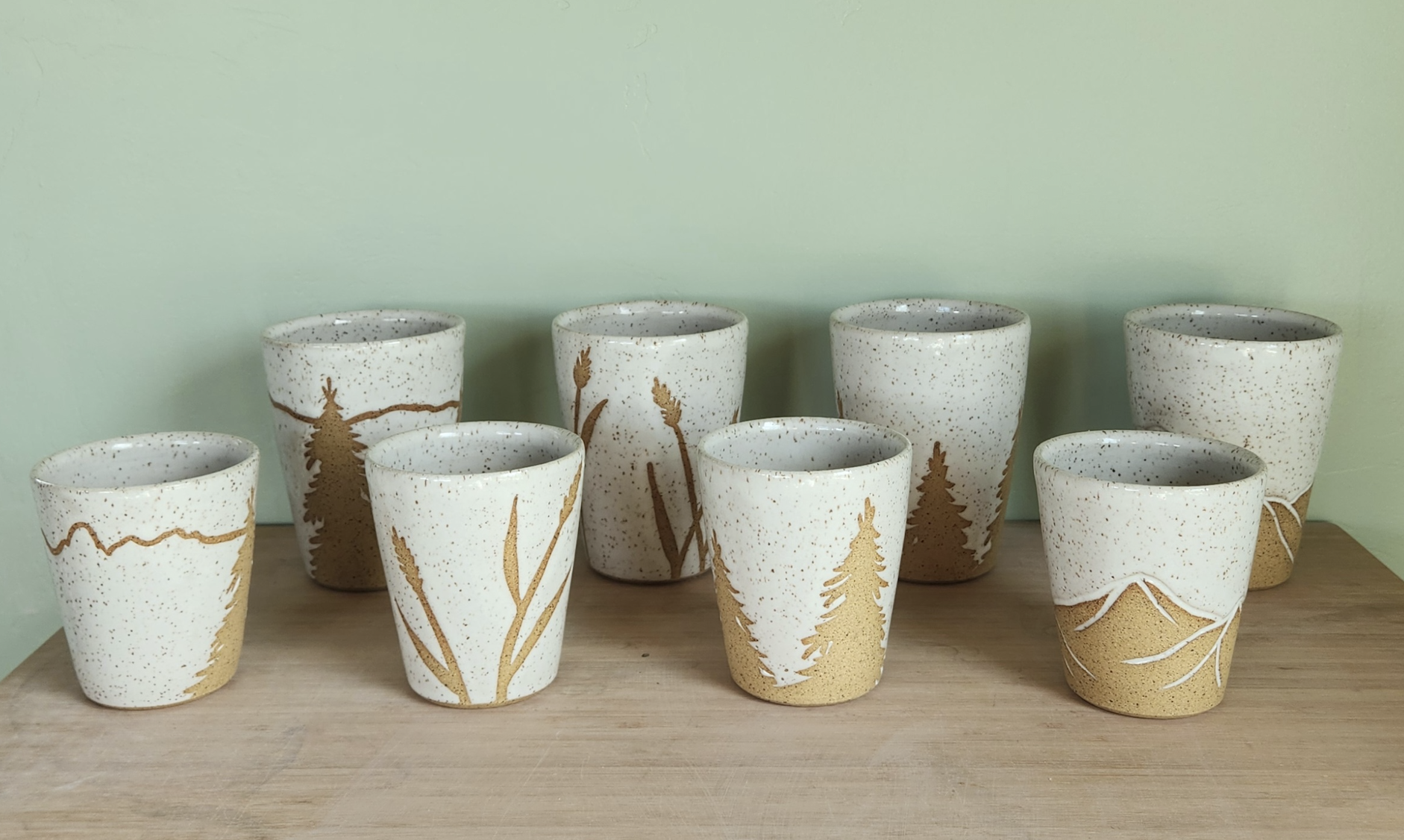 Set of eight ceramic cups with a nature theme, featuring gold silhouette of trees and plants, arranged on a wooden surface against a pale green wall.