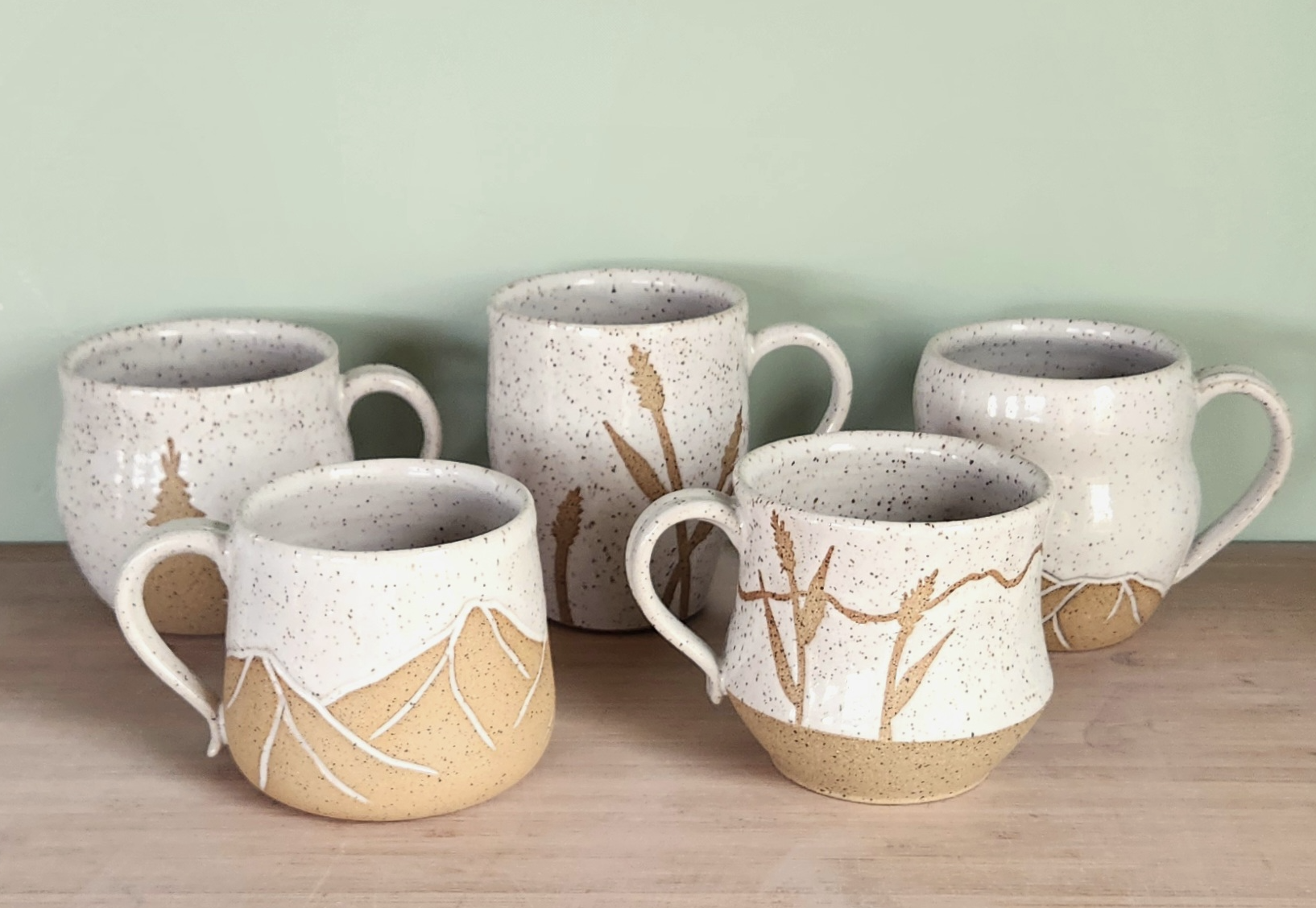 Five speckled beige ceramic mugs with nature-inspired designs of branches and plants, arranged on a wooden surface against a light green wall.