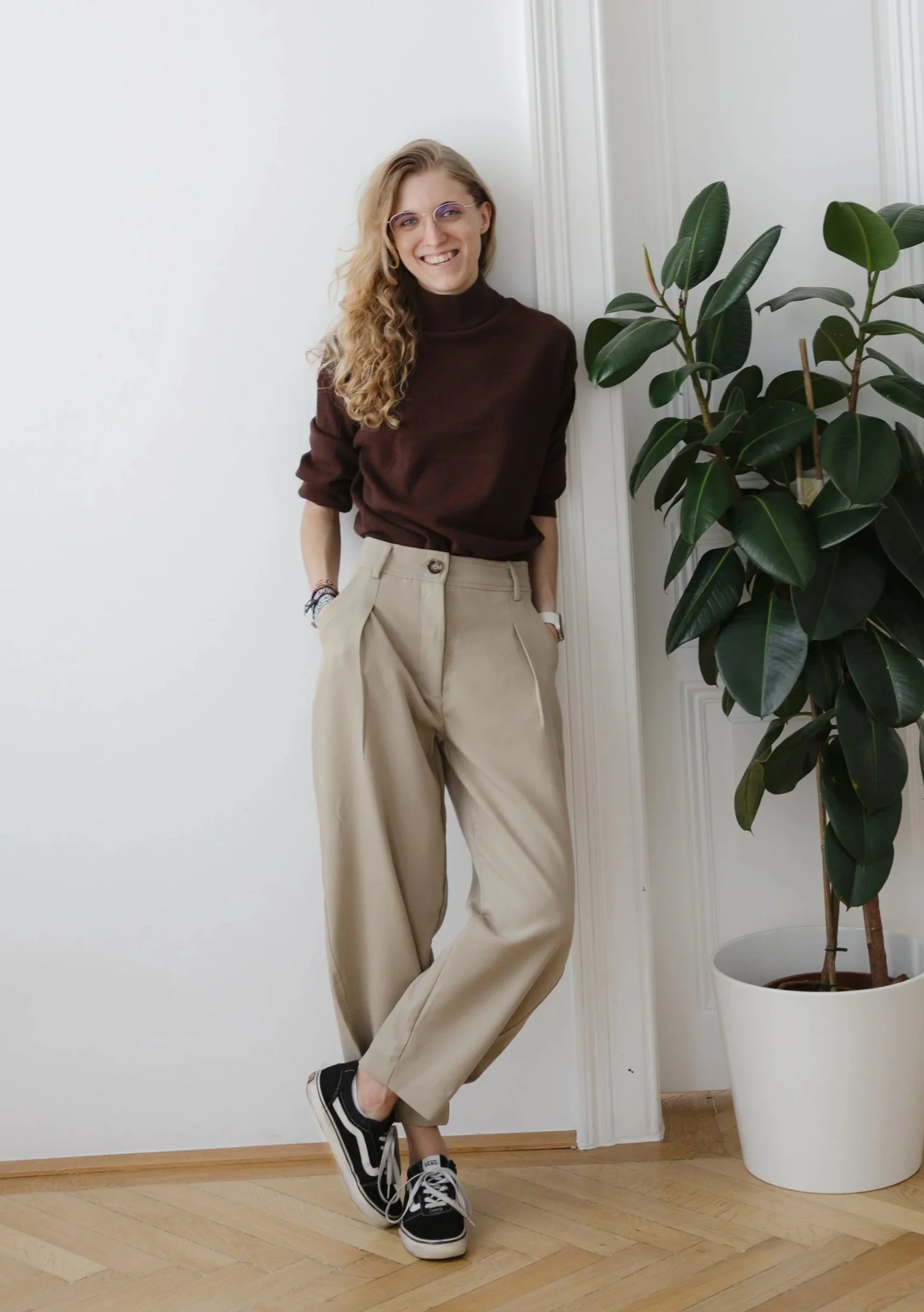 Celina Klestorfer, Designerin für Websites und Branding - eine blonde Frau in dunkelbraunem Pullover und beige Hose lehnt an einer weißen Wand.