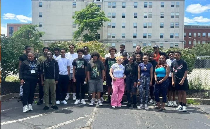 Group of diverse young people standing in a parking lot outdoors with a building and trees in the background.