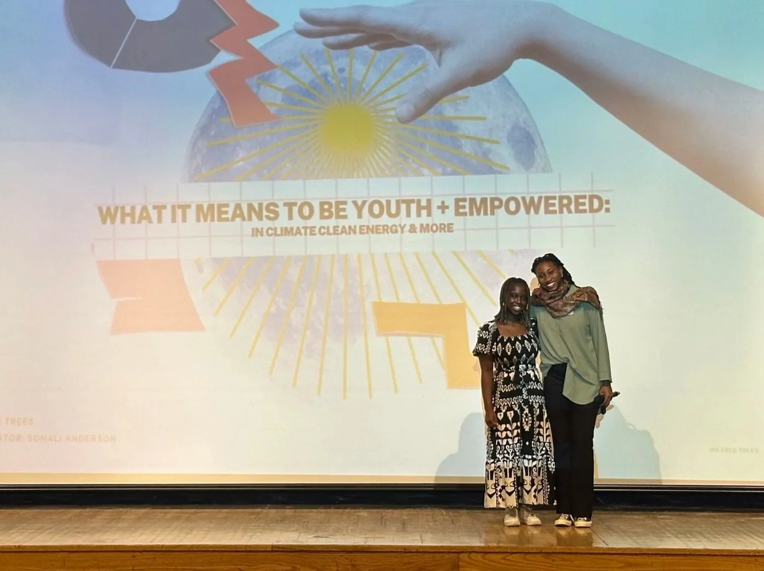 Two women standing on a stage in front of a large presentation slide. The slide has a graphic of the moon with a hand reaching toward it and colorful rays emanating from it. There is text on the slide that reads, "WHAT IT MEANS TO BE YOUTH + EMPOWERED: IN CLIMATE CLEAN ENERGY & MORE." The women are smiling, one wearing a black and white patterned dress and the other wearing a green top with a scarf and black pants.