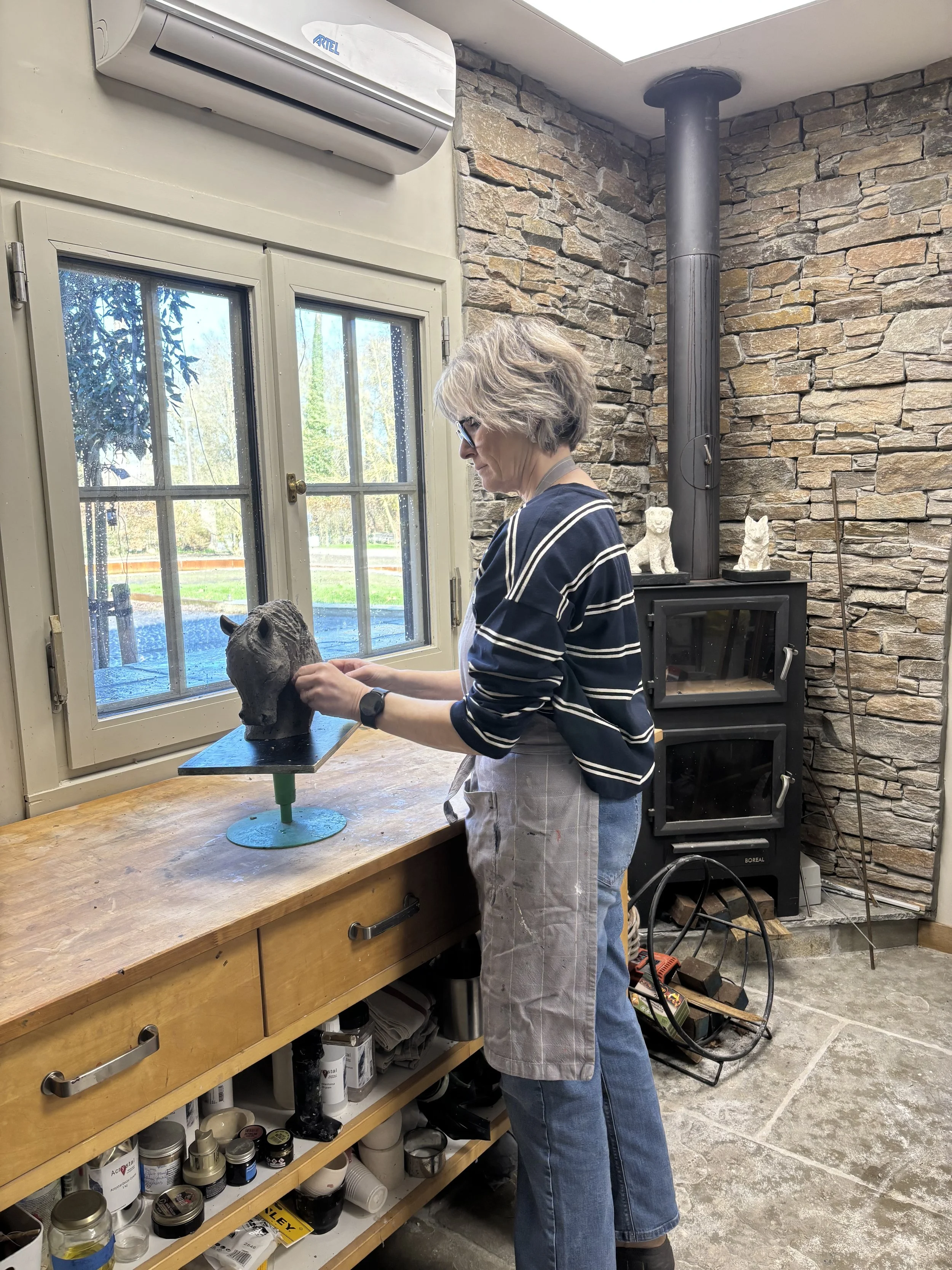 Middeloude vrouw in een werkbroek en een gestreept shirt die een beeld van een paard bewerkt in een kunstatelier met een stenen muur, een houtkachel en een raam met uitzicht op een tuin.