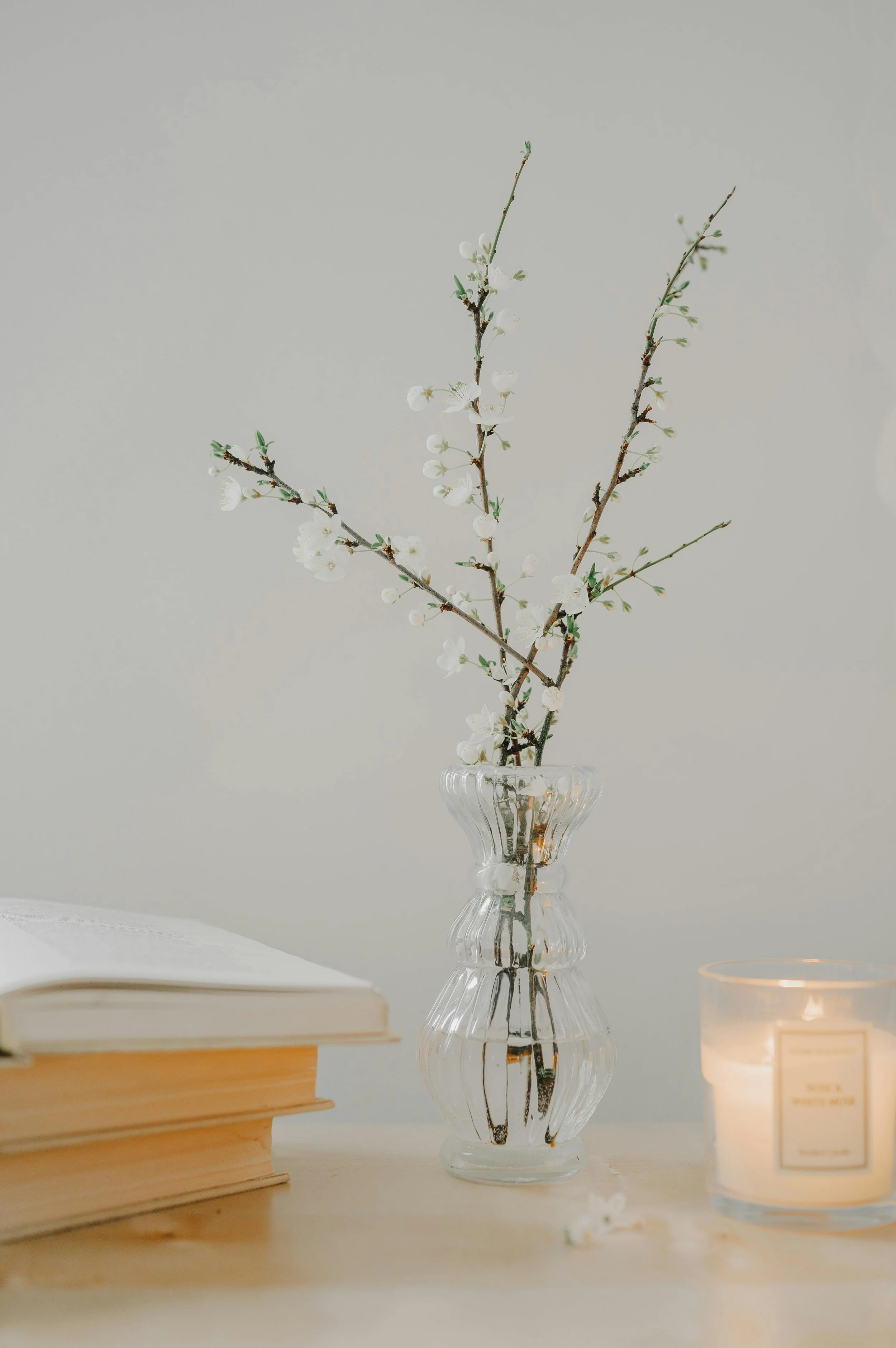 A clear glass vase with white flowering branches, an open book, and a lit candle on a wooden surface against a plain background.