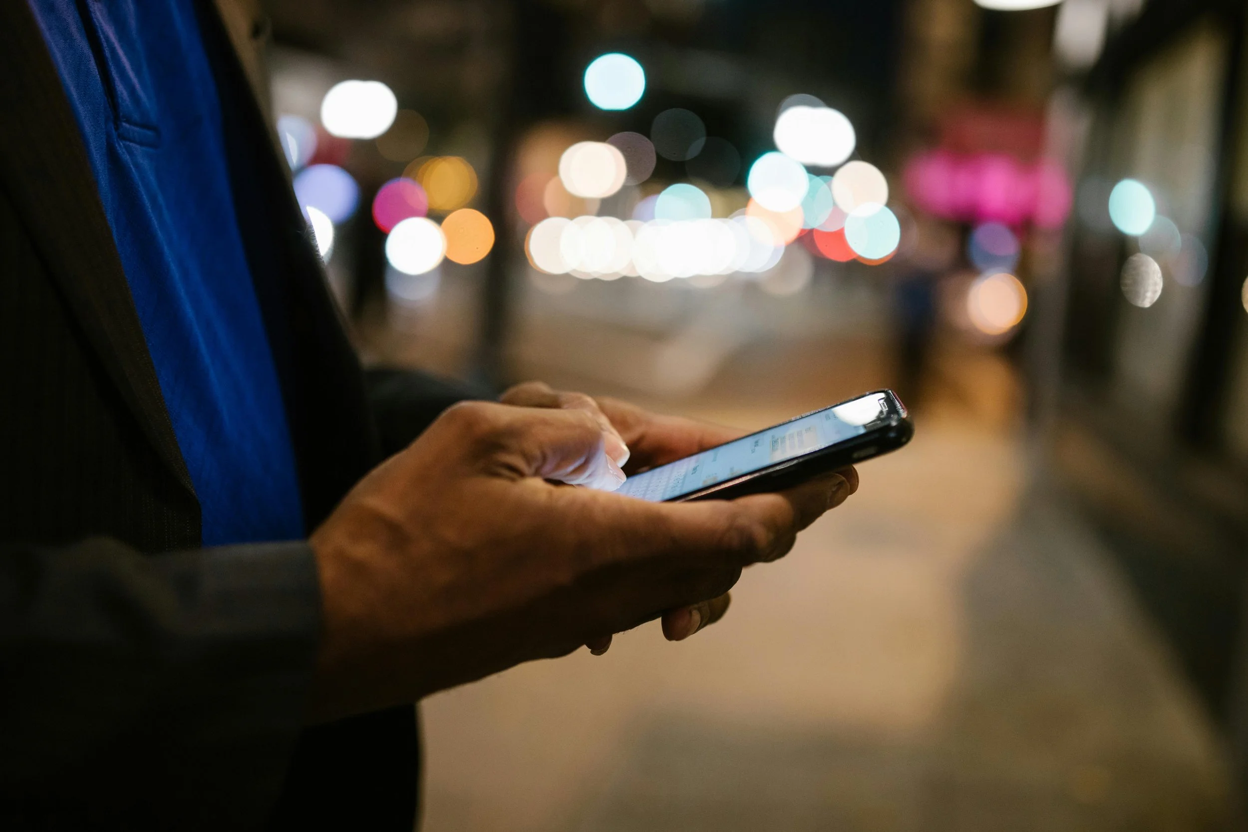 Person using a smartphone on a city street at night, with blurred colorful lights in the background.