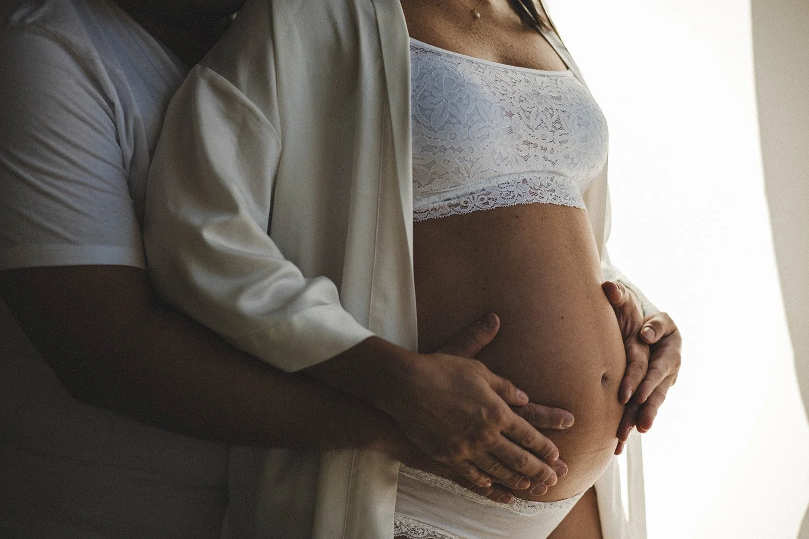 Pregnancy portrait of a woman standing by a window in natural light during an intimate maternity session.