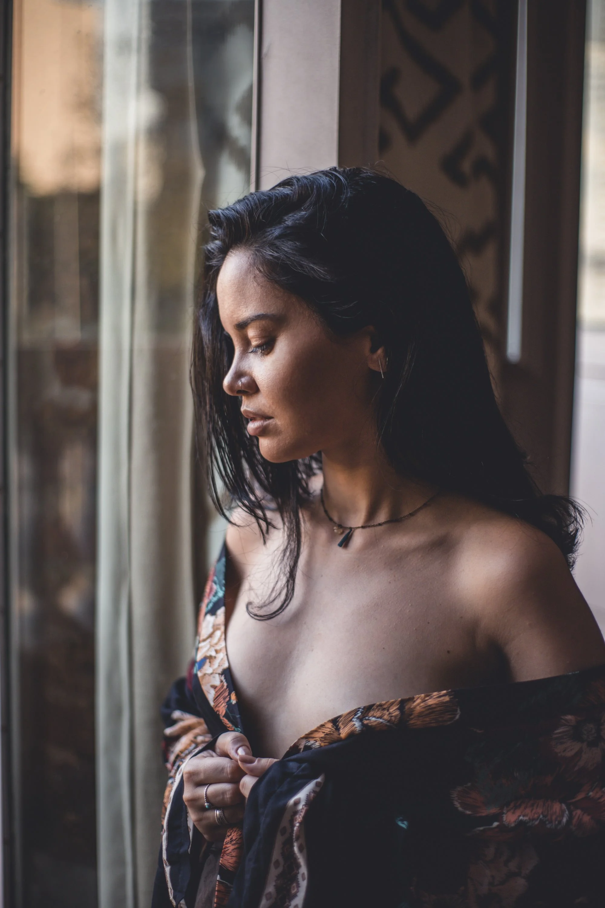 Intimate portrait of a woman standing by a window in soft natural light during a boudoir photoshoot.