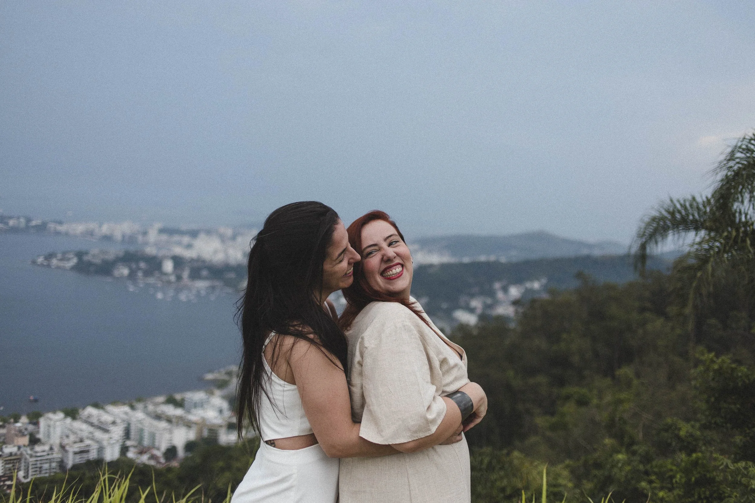 A lesbian couple embracing during an engagement photo session with a scenic city view of Rio de Janeiro in the background.