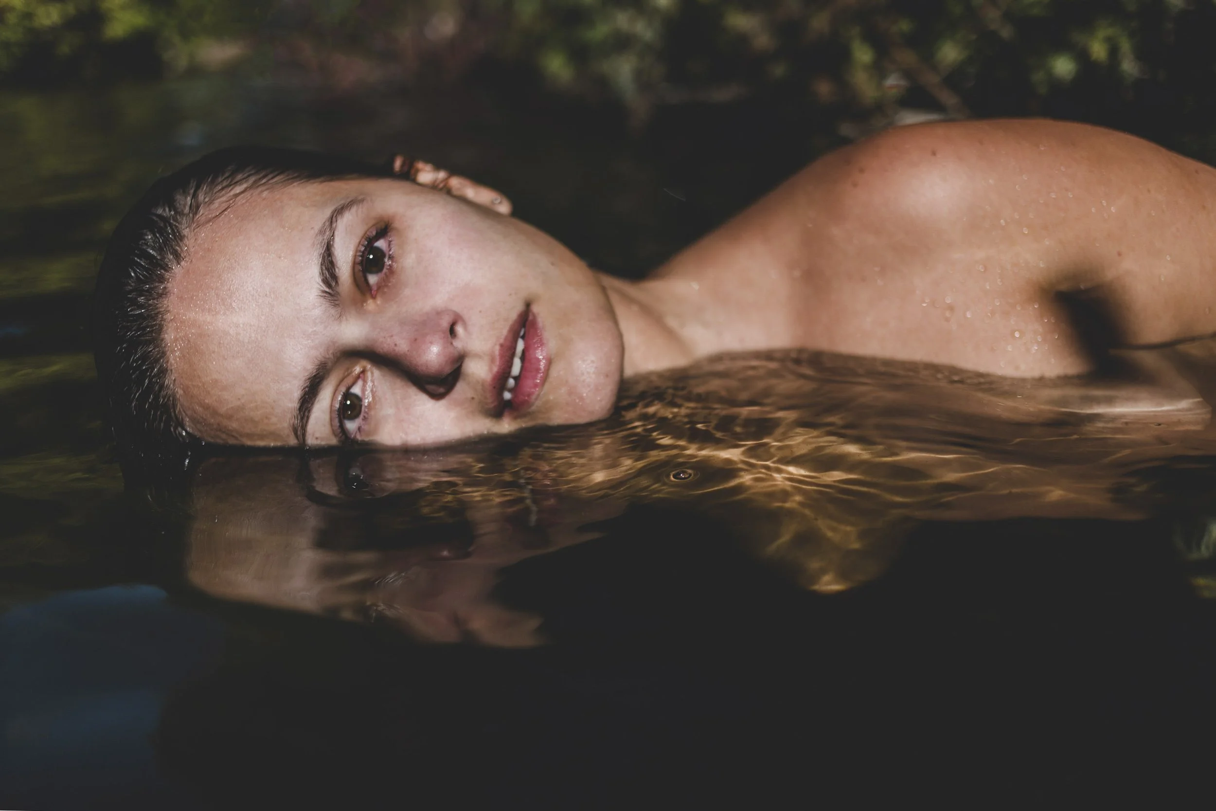 Boudoir portrait of a woman photographed in nature, with wet hair and a calm, reflective expression.
