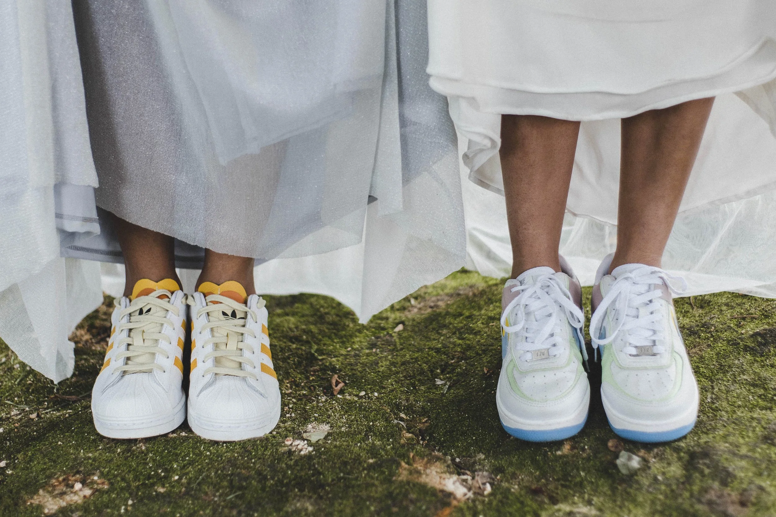 Detail shot of a lesbian couple during a wedding photo session, showing their shoes and dresses standing together on mossy ground.