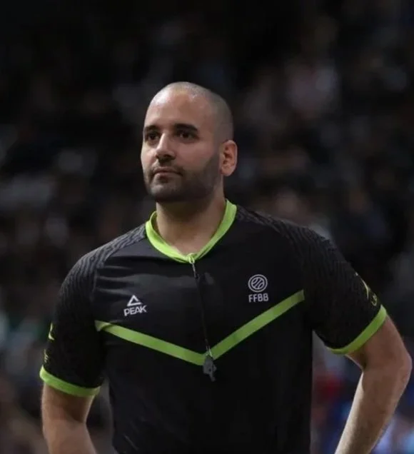 Un arbitre de football portant un maillot noir avec des détails verts, avec un chronomètre autour du cou, dans un stade rempli de spectateurs.