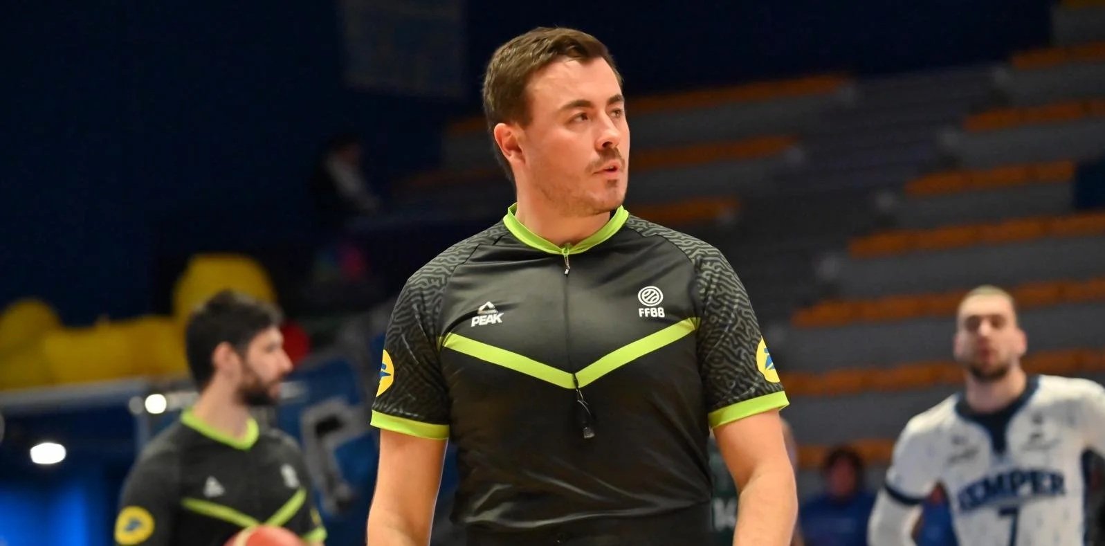 Arbitre de rugby en tenue noire avec des accents jaunes, dans une salle de sport ou stade, avec deux joueurs en arrière-plan.