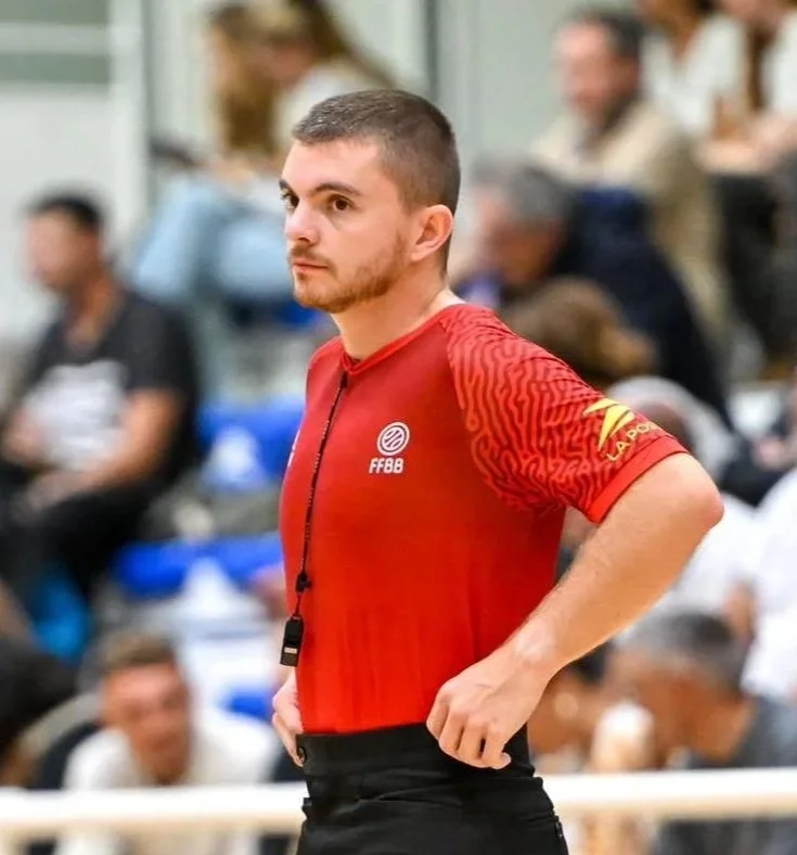 Un arbitre ou entraîneur portant un maillot rouge avec le logo de la fédération française de basket-ball (FFBB), pendant un match en salle avec des spectateurs en arrière-plan.
