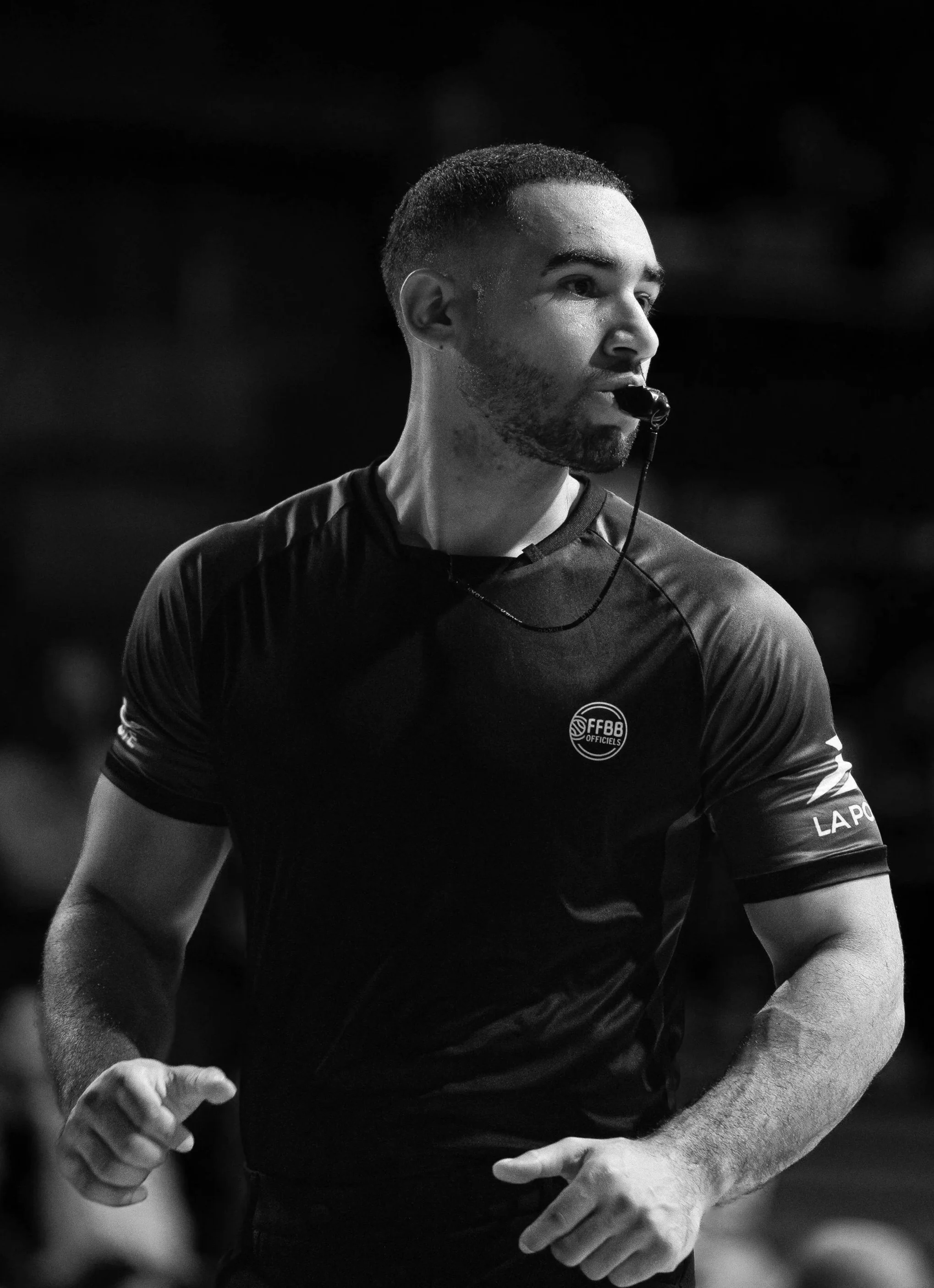 Arbitre en action lors d'un match, portant un maillot avec le logo de la Fédération Française de Basketball, en noir et blanc.