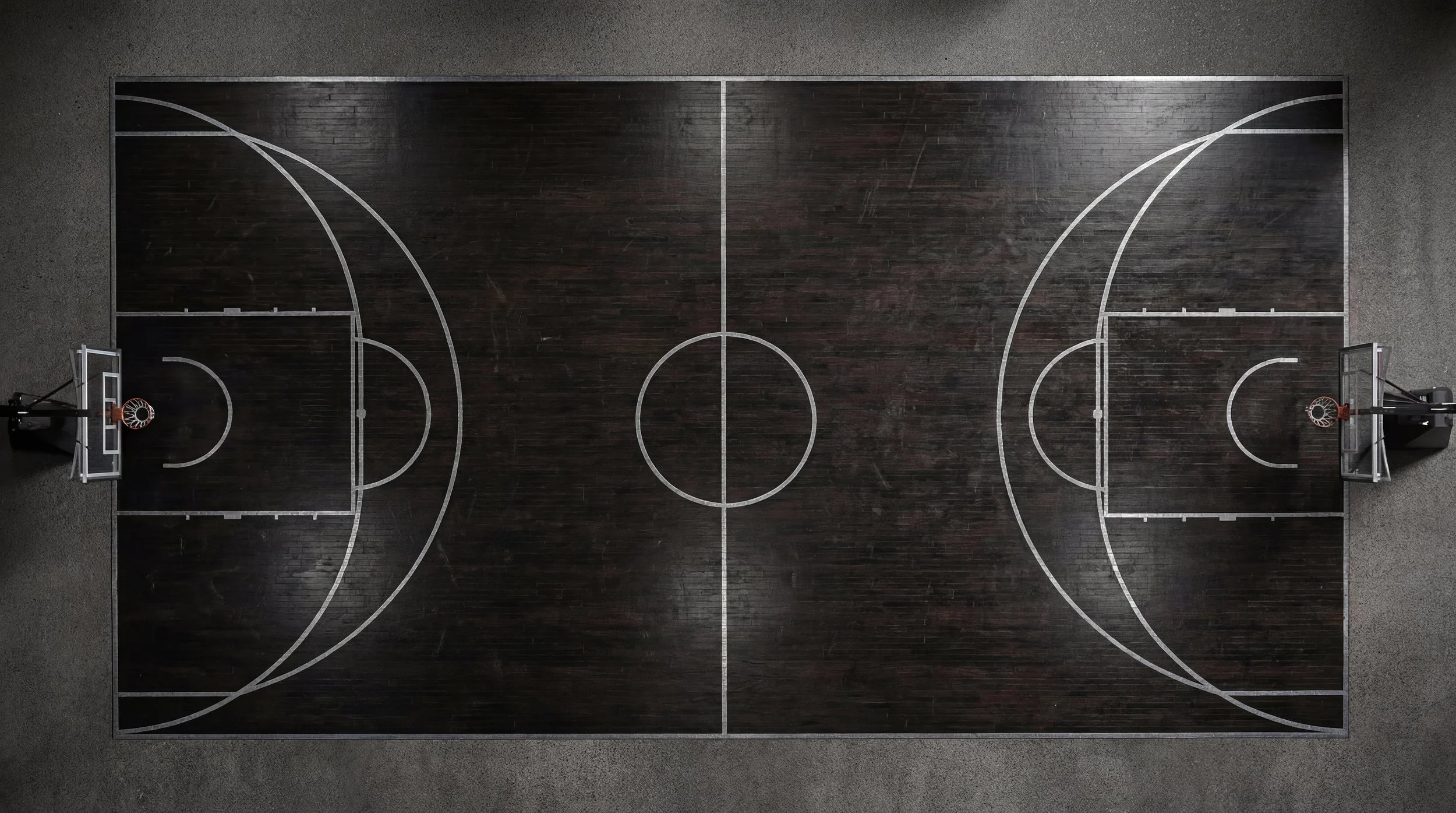 Vue aérienne d'un terrain de basketball en bois sombre avec lignes blanches, deux paniers de basket à chaque extrémité, et un sol en béton gris environnant.