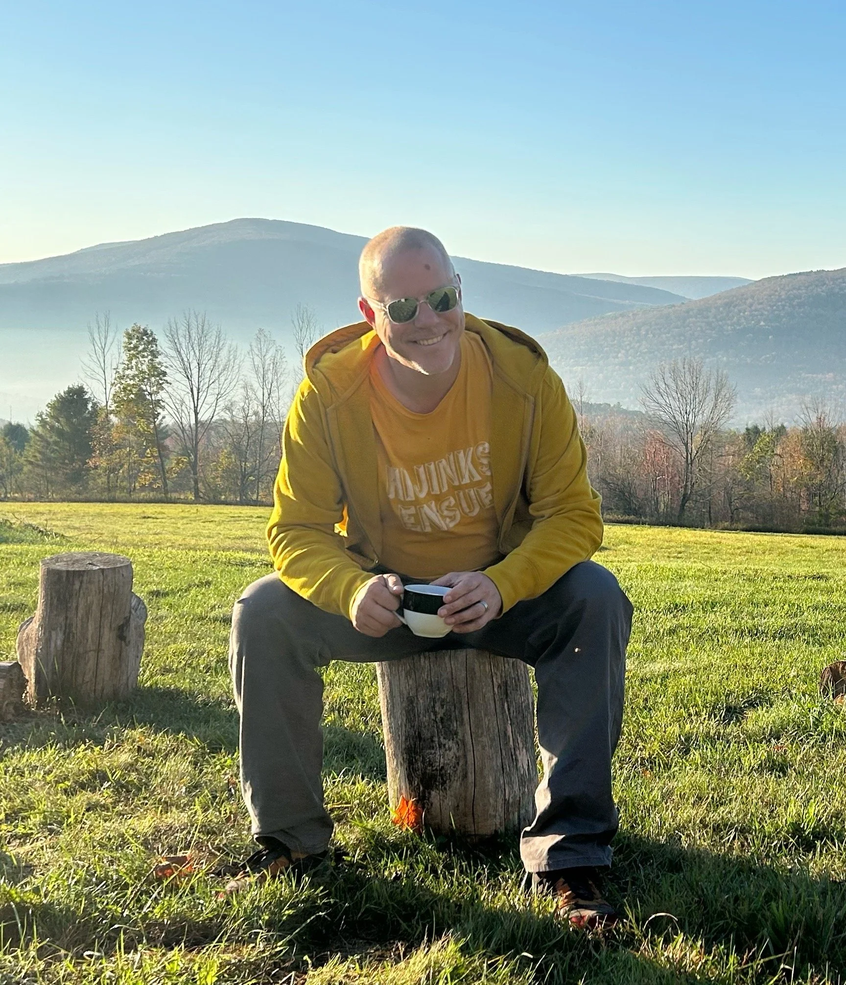 Tim sitting on a stump, holding a coffee cup and smiling, with a view of rolling hills in the morning in the background.