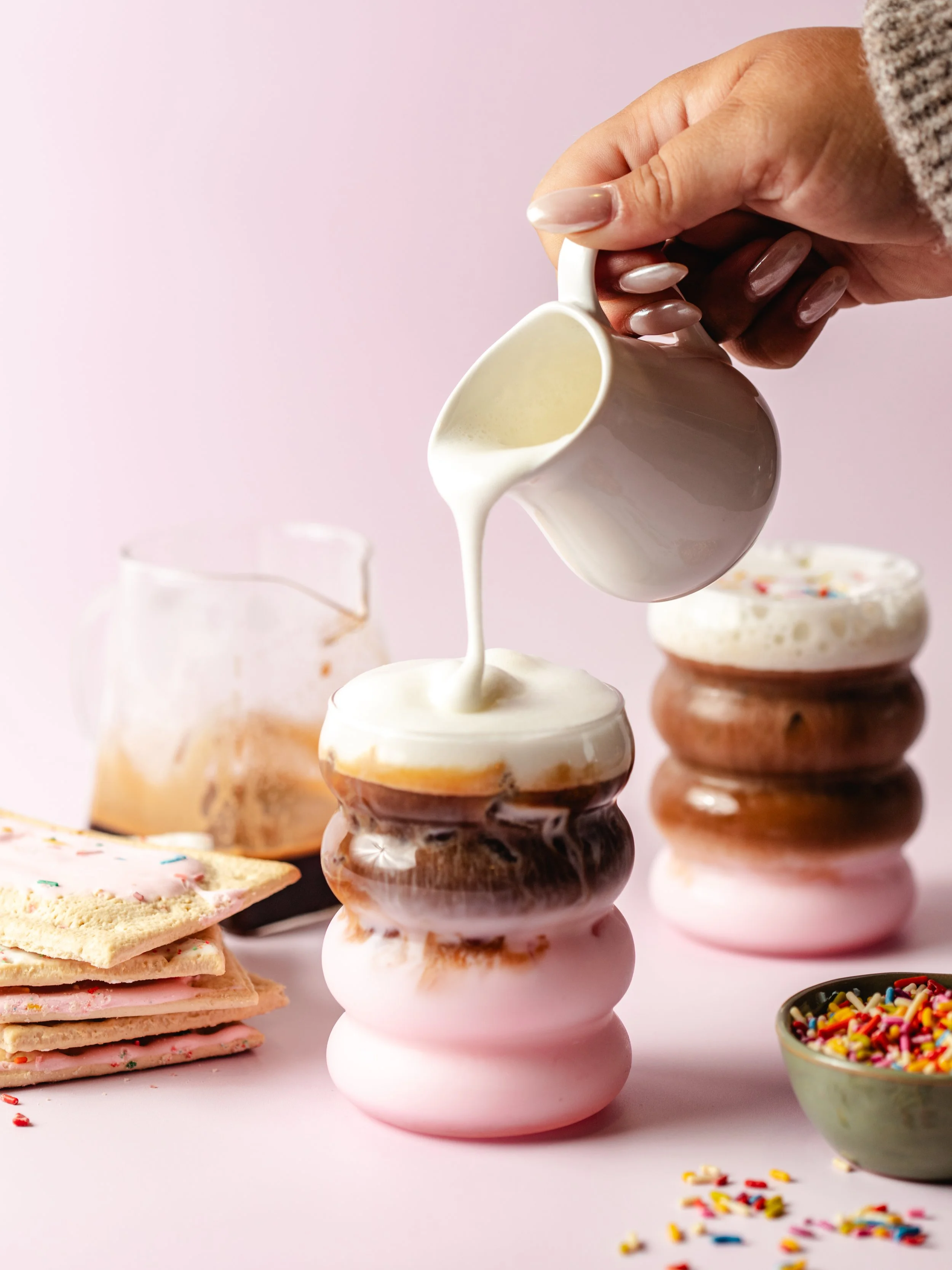A person pouring milk onto a stack of pink, brown, and white donuts with frosting, sprinkles, and cookies, with additional donuts and toppings in the background on a pink surface.