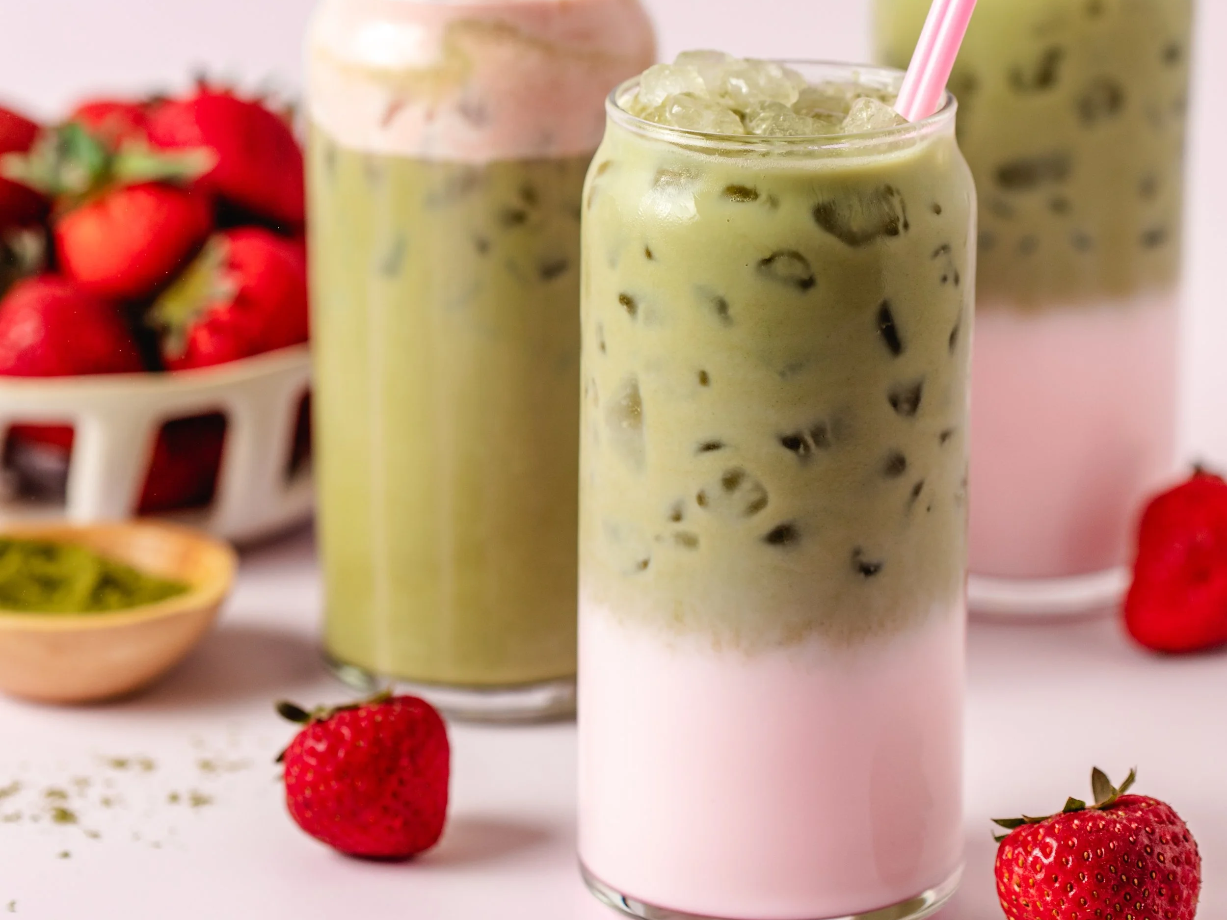 Three iced matcha drinks with pink, green, and white layers in tall glasses on a white surface, with strawberries and a small bowl of passion fruit around them.