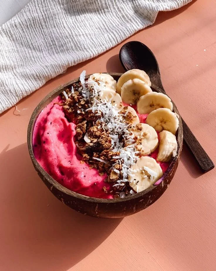 A bowl of pink smoothie topped with banana slices, granola, shredded coconut, and a swirl of pink berry sauce, with a black spoon beside it on a light pink surface with a white cloth nearby.