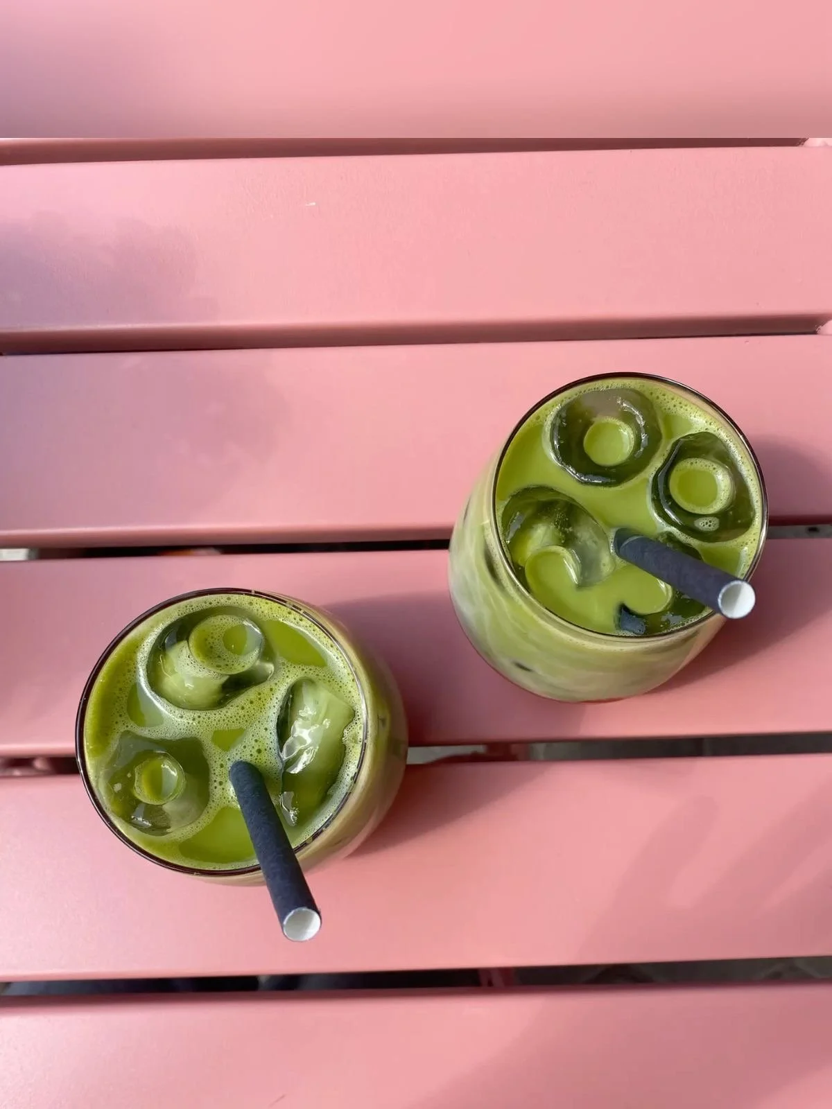 Two glasses of iced green matcha latte on a pink wooden table with black straws.
